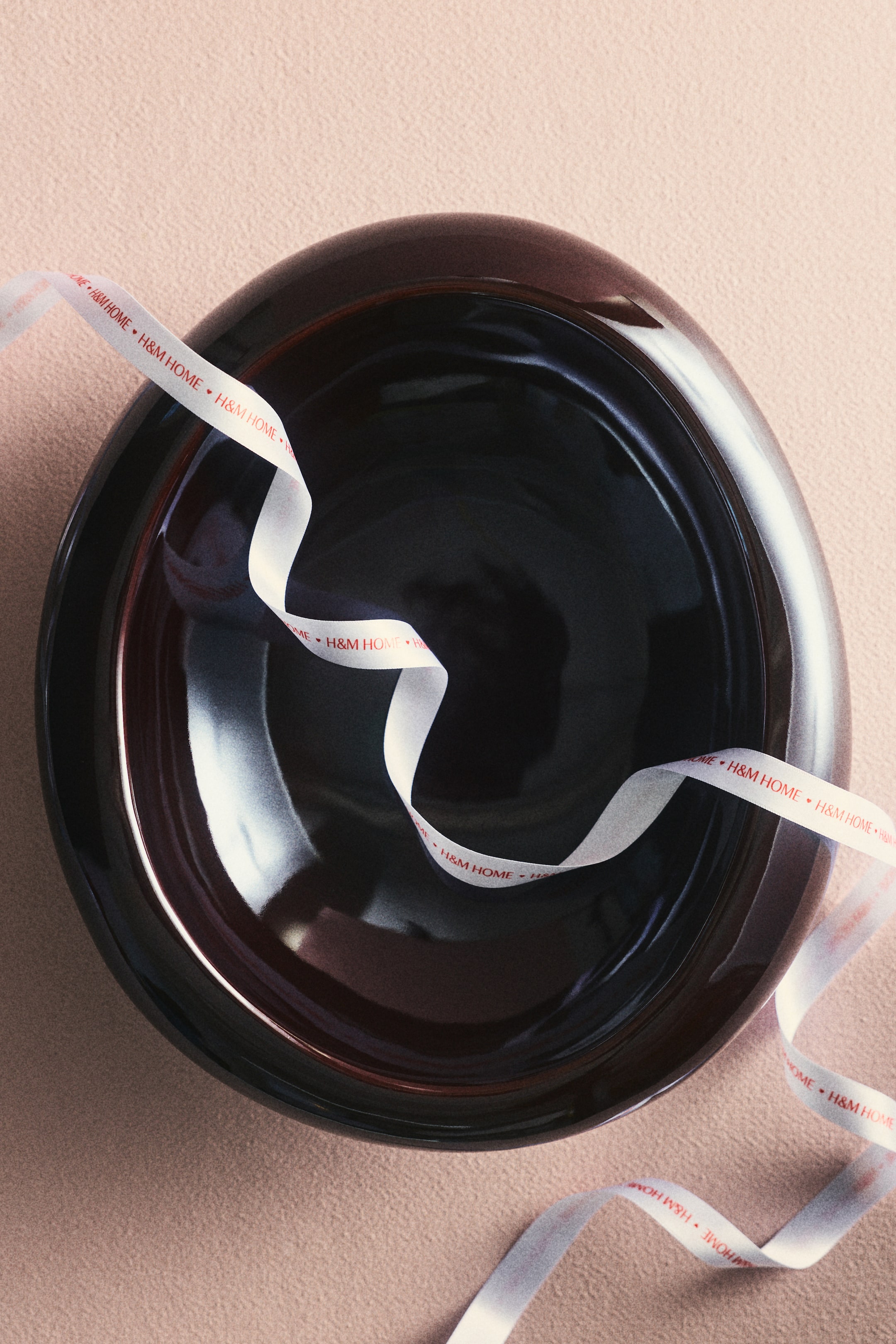 Zobrazit větší obrázek: Tmavě hnědá, vysoce lesklá mísa zobrazená zhora má kruhový tvar s prohloubeným vnitřkem a výrazným vystouplým okrajem. Bílá stužka s potiskem "H&M HOME" se volně přehýbá přes její povrch a leží uvnitř mísy.
