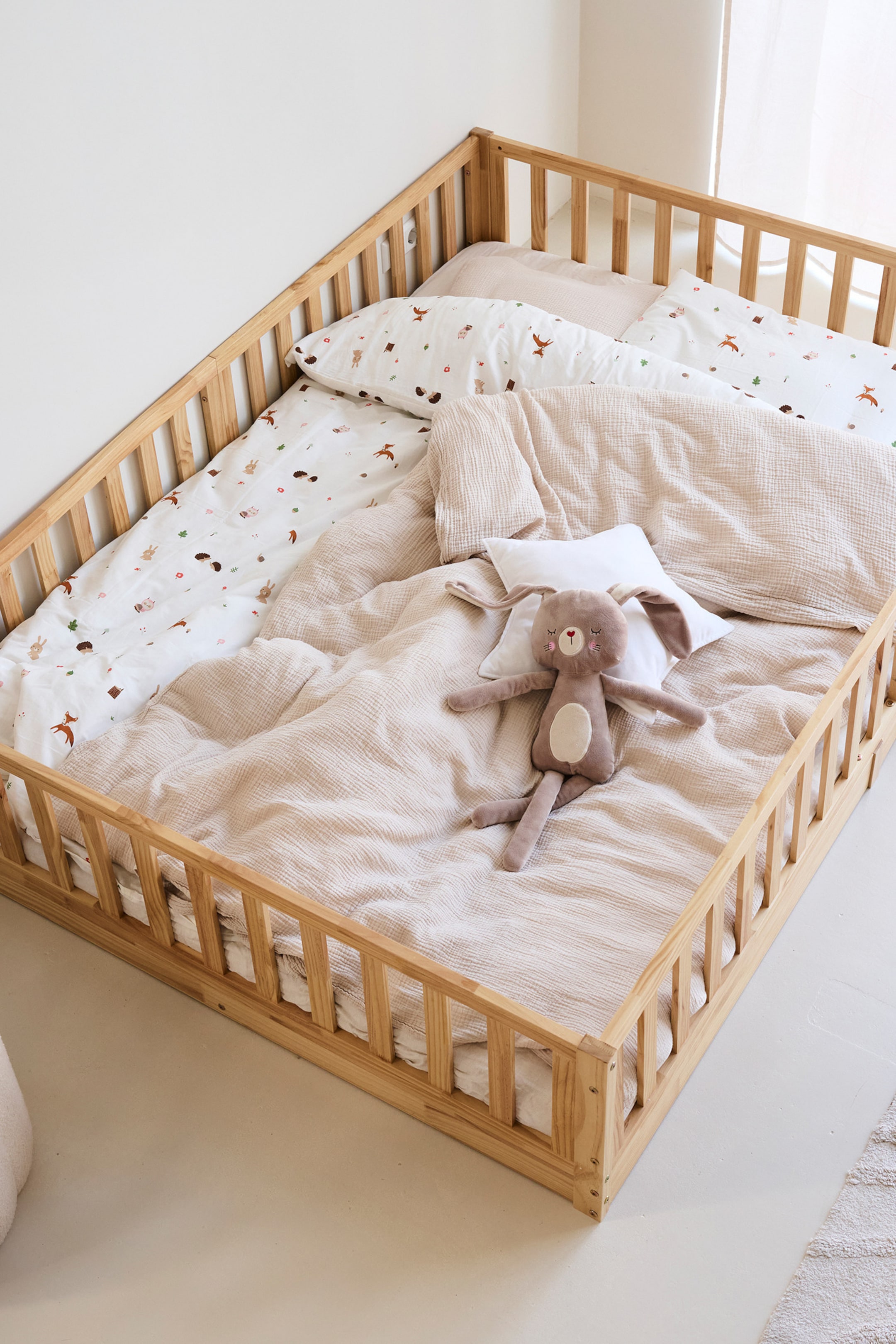 Agrandir l'image: Un cadre de lit au sol en bois naturel est présenté avec des côtés à lattes vus d'en haut. À l'intérieur, une couette et un oreiller blancs arborent un motif d'animaux de la forêt, tandis qu'une couverture froissée beige clair et un jouet en peluche en forme de lapin brun sont également présents.
