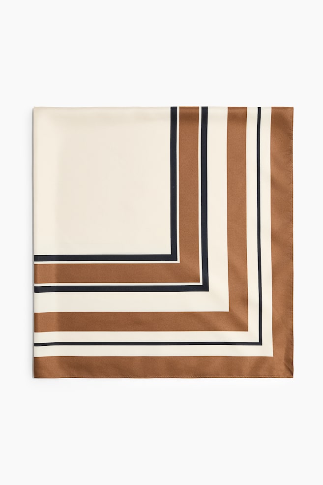Ein quadratisches Tuch in weicher Webart liegt flach und mit der Vorderseite nach oben und zeigt ein gedrucktes geometrisches Muster. Das Design weist in einer Ecke einen einfarbigen cremefarbenen Block auf, der von abwechselnden Streifen in Braun, Marineblau und Creme eingefasst ist. - 1