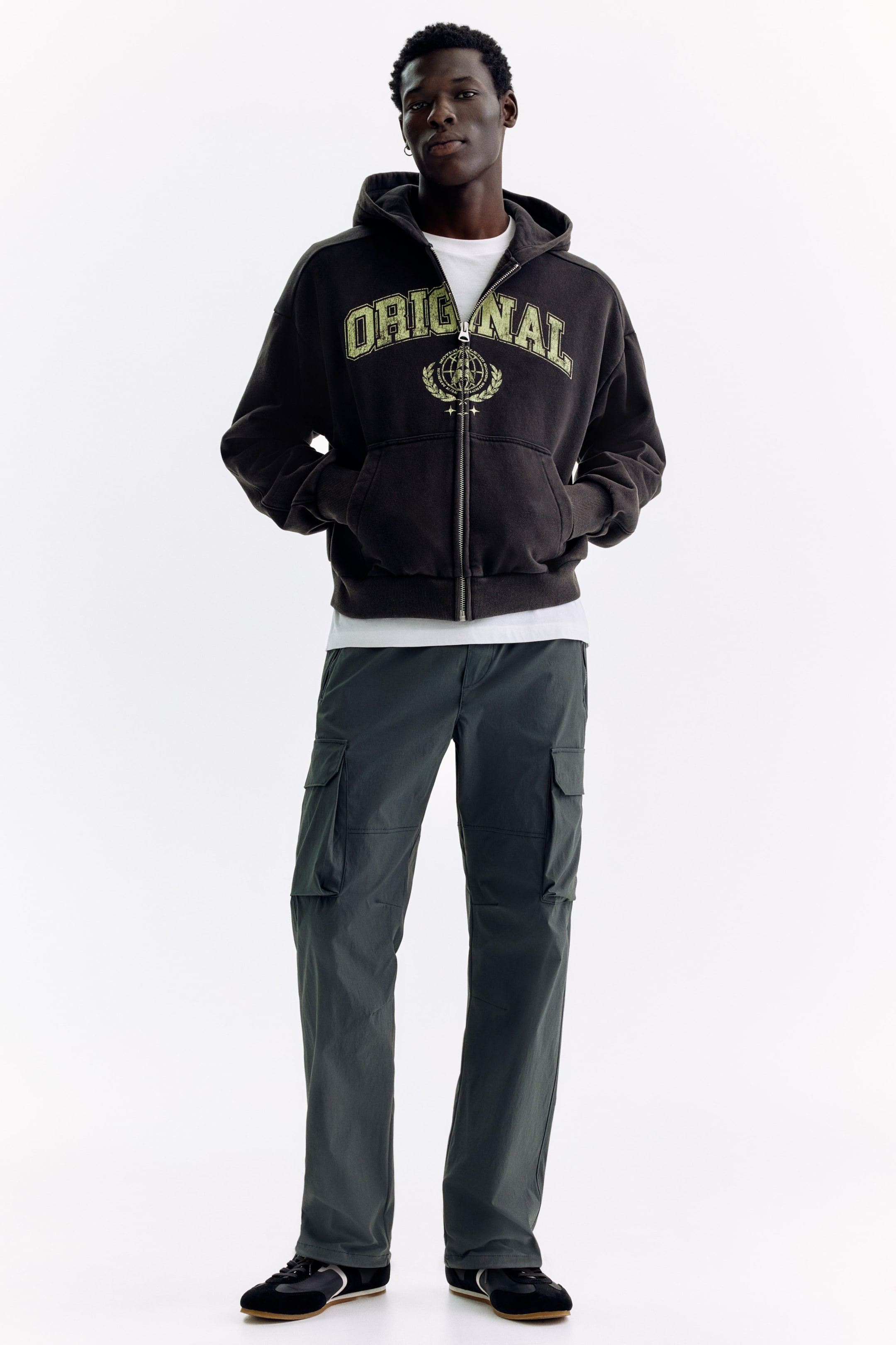 Voir une image plus grande: Le chandail à capuchon zippé brun foncé délavé est orienté vers l'avant, arborant un graphique "ORIGINAL" vert pâle vieilli et un emblème sur la poitrine. Il présente une fermeture éclair pleine longueur, une poche kangourou et des poignets et ourlet côtelés, avec un t-shirt blanc en dessous.