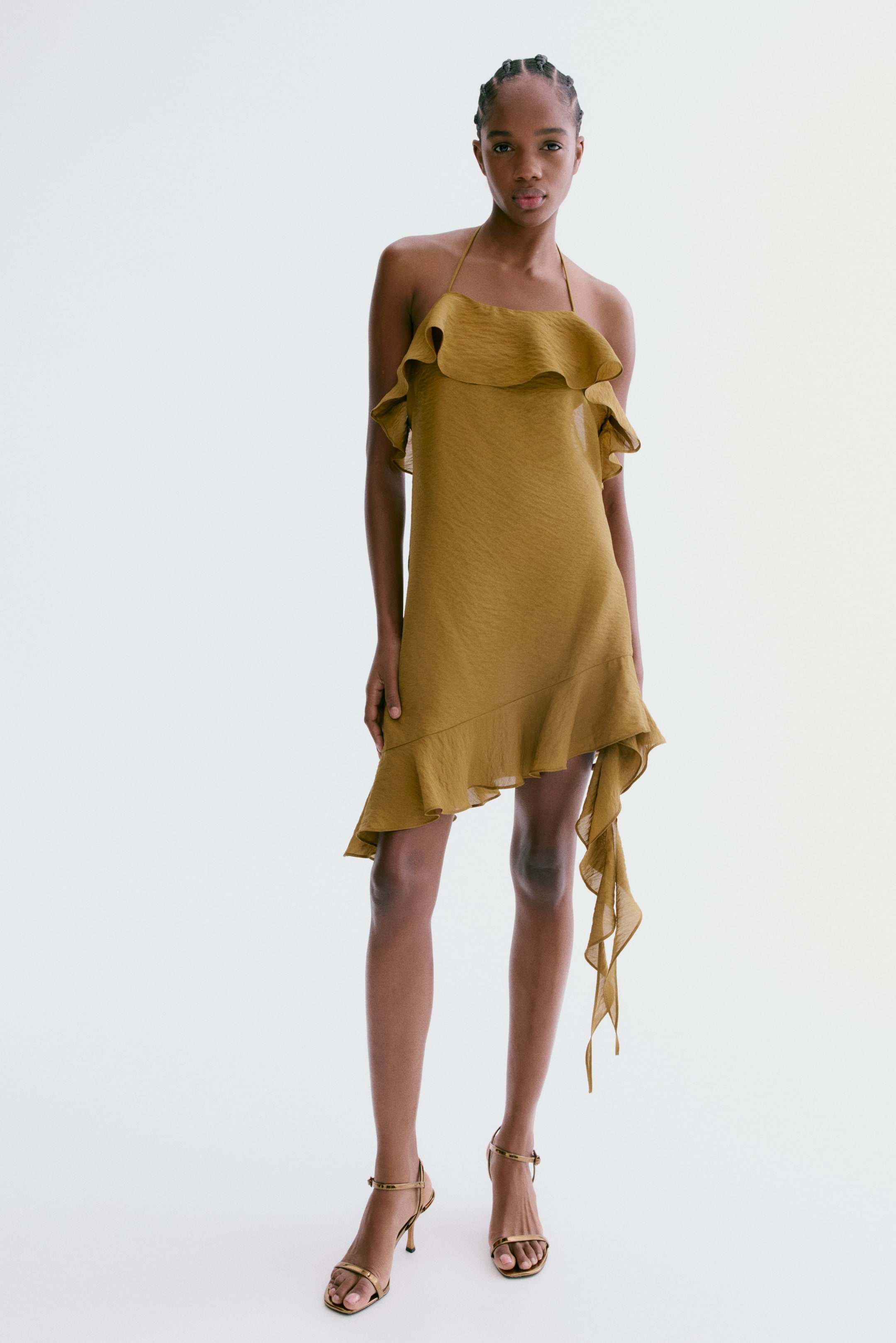 View larger image: A golden brown textured mini dress featuring a halter neck and a ruffled overlay across the bust. The dress showcases an asymmetrical hemline with a cascading ruffle extending longer on one side, paired with metallic gold strappy stiletto sandals.