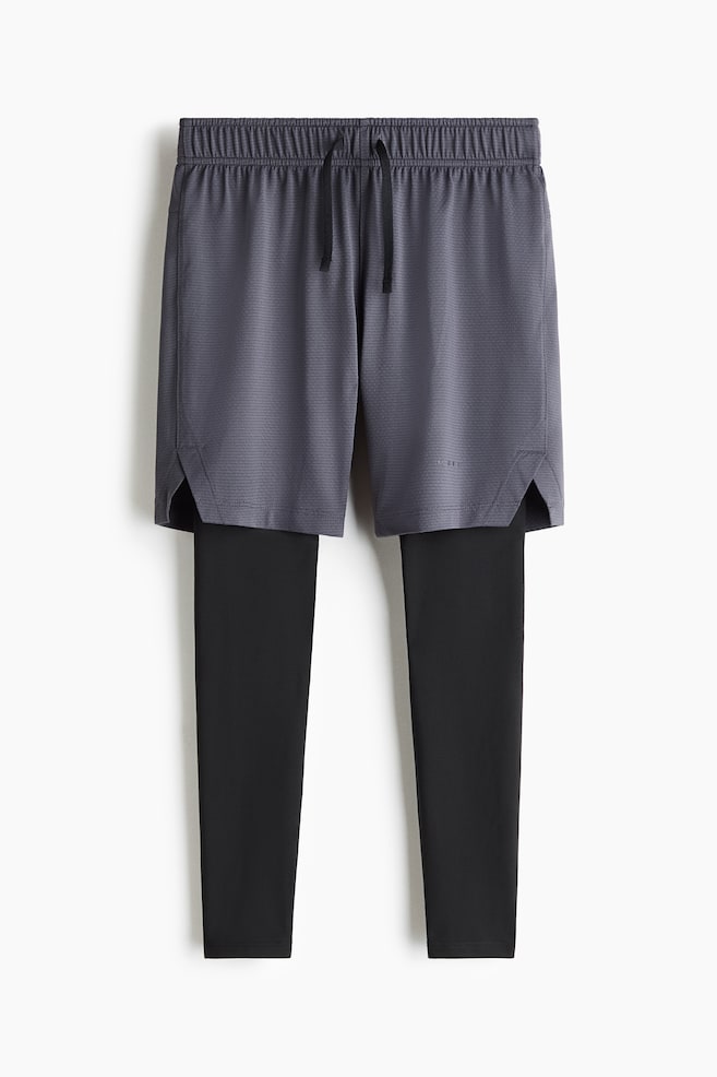 Un par de pantalones deportivos dos en uno presenta shorts texturizados grises superpuestos sobre leggings negros integrados, mostrados de frente. Los shorts exteriores muestran una cintura elÃ¡stica con cordÃ³n negro y dobladillos laterales con abertura. - 1