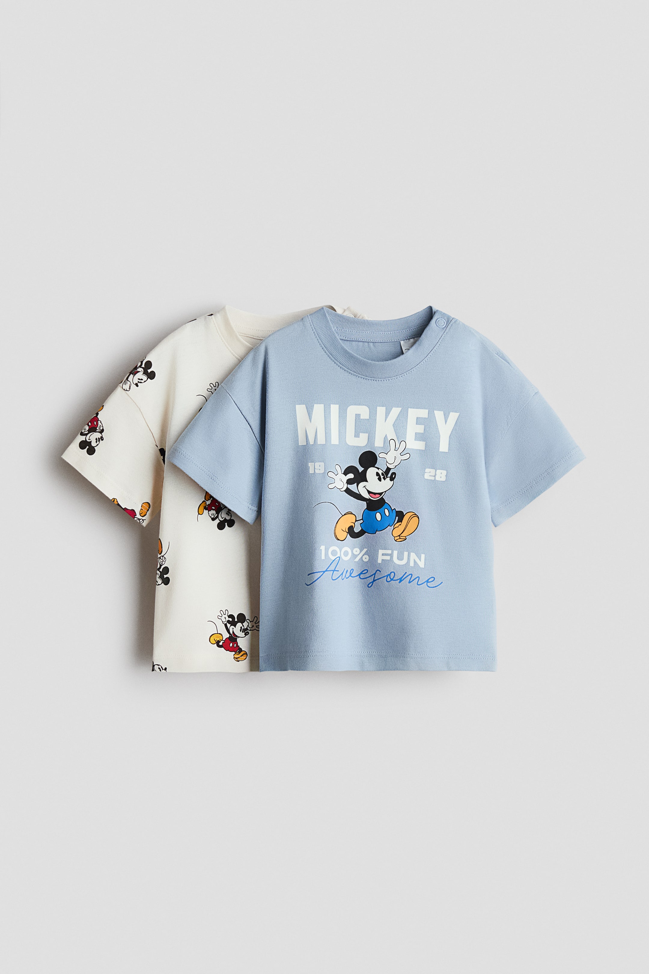 View larger image: Two baby T-shirts are presented facing forward, with a light blue T-shirt in the foreground featuring a large Mickey Mouse graphic, "MICKEY" text, and "100% FUN Awesome" lettering, plus two snap buttons on the left shoulder. A cream-coloured T-shirt is layered behind it, displaying a repeating pattern of smaller Mickey Mouse figures.