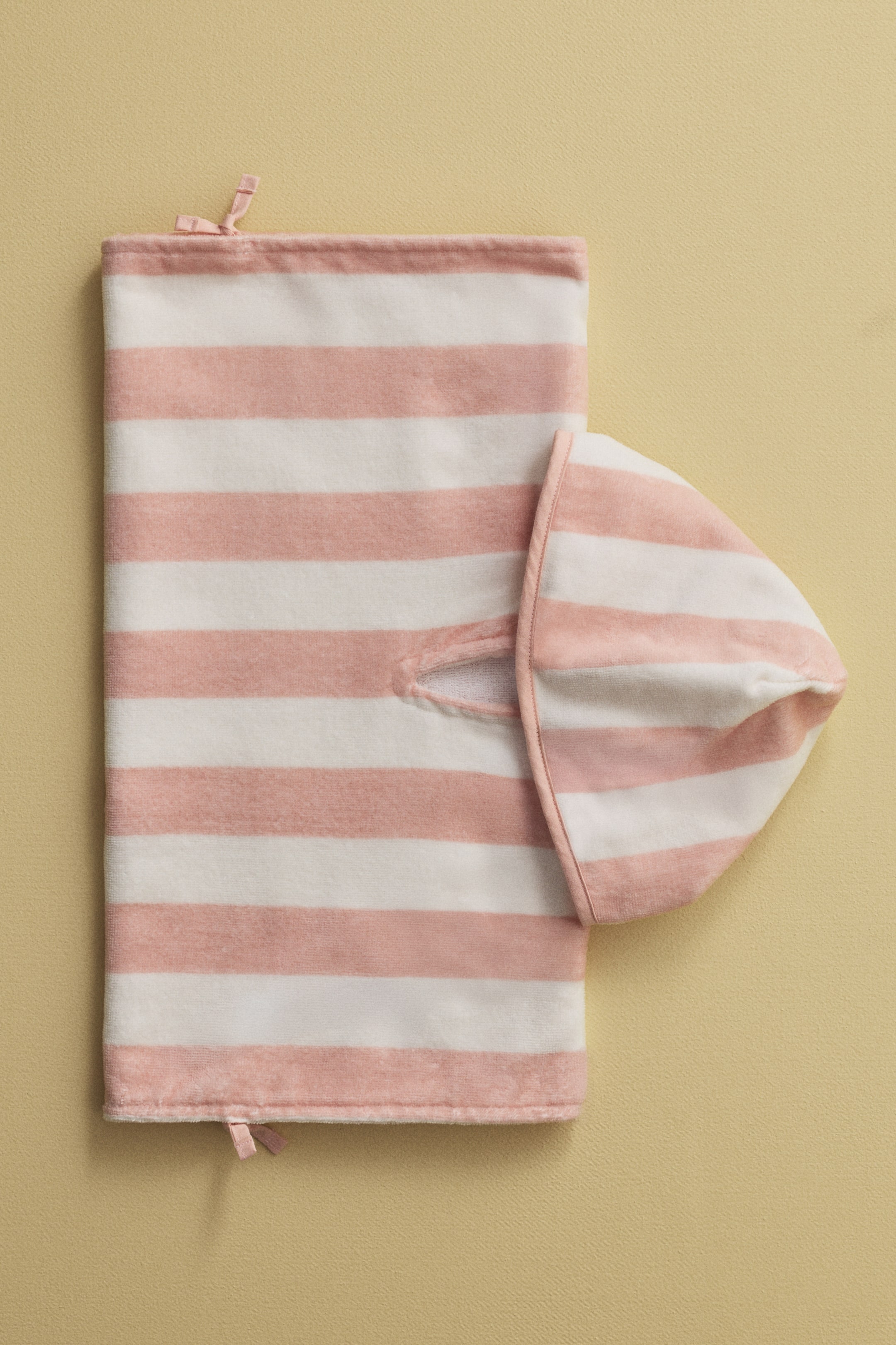 Nagyobb kép megtekintése: Egy rózsaszín és fehér csíkos poncsó törölköző elölről mutatkozik, laposan elhelyezve, puha, velúr-szerű textúrával, vízszintes csíkokkal. Egy csatolt kapucni látható, részben felfedett nyílással, és keskeny rózsaszín kötők jelennek meg az oldalsó széleken.