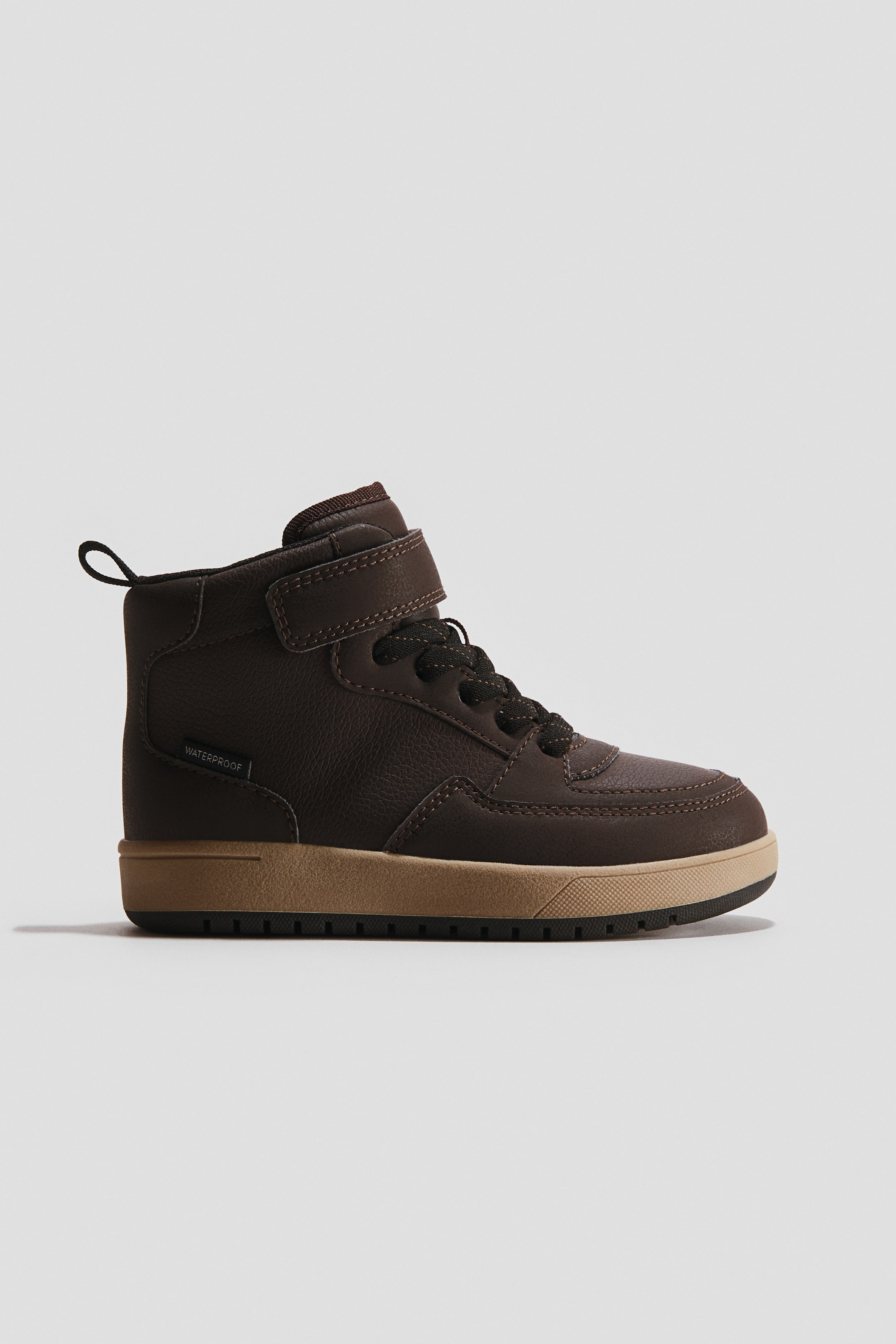 Se større billede: En mørkebrun høj sneaker vender mod højre og viser sin sideprofil. Den har en struktureret overdel med synlige syninger, sorte og brune spættede snørebånd, en mørkebrun velcrostrop, der sikrer anklen, og en sort trækstrop, der strækker sig fra bagsiden. "WATERPROOF" er trykt på siden, og en tan mellemsål står i kontrast til en sort, rillet ydersål.