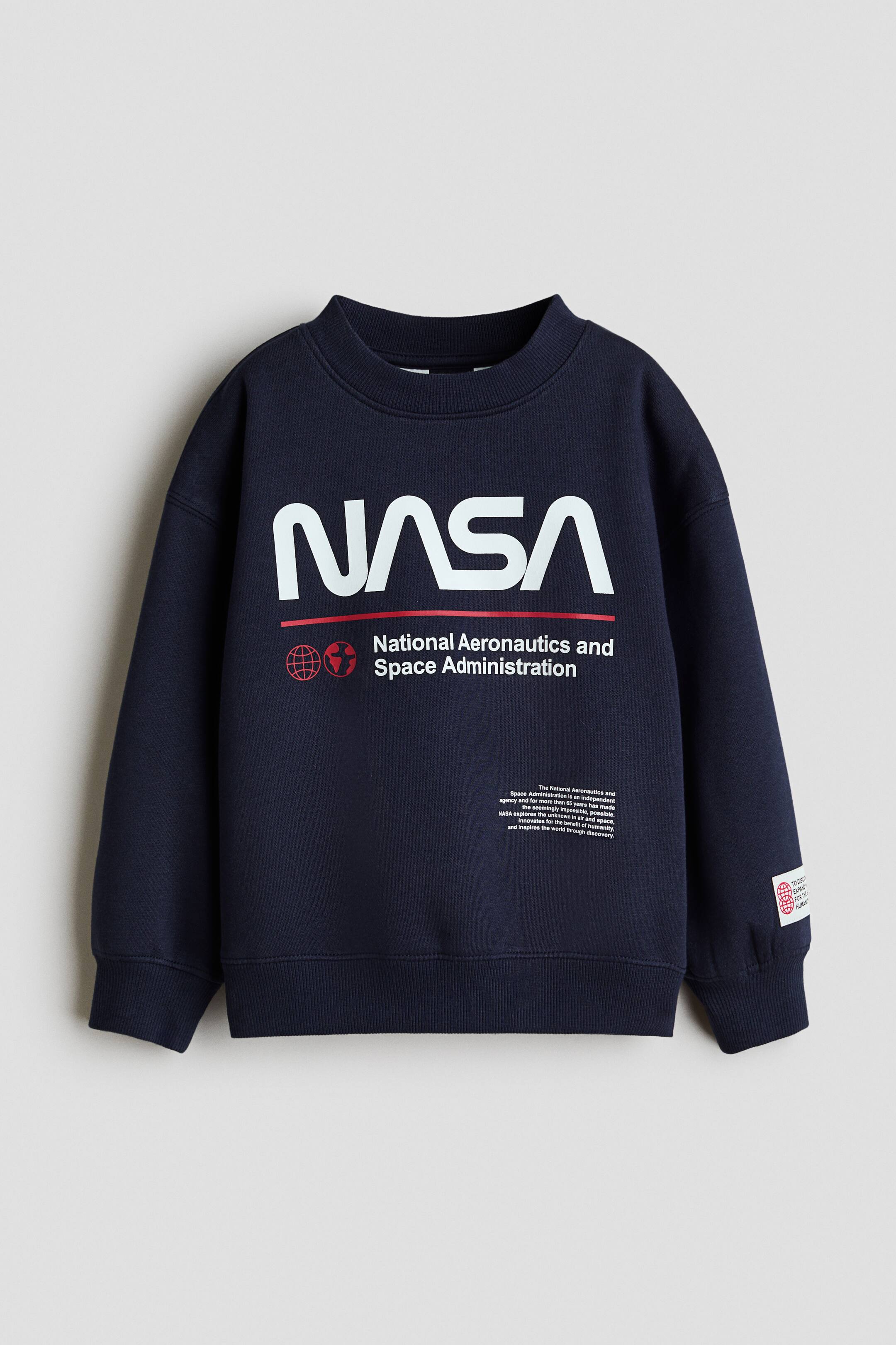 Visa större bild: En marinblå sweatshirt visas framifrån och har en stor vit "NASA"-logotyp med ett rött understreck och text tryckt över bröstet. Plagget med lös passform har nedhasade axlar, ribbad rund hals och ribbade muddar vid ärmslut och nederkant.