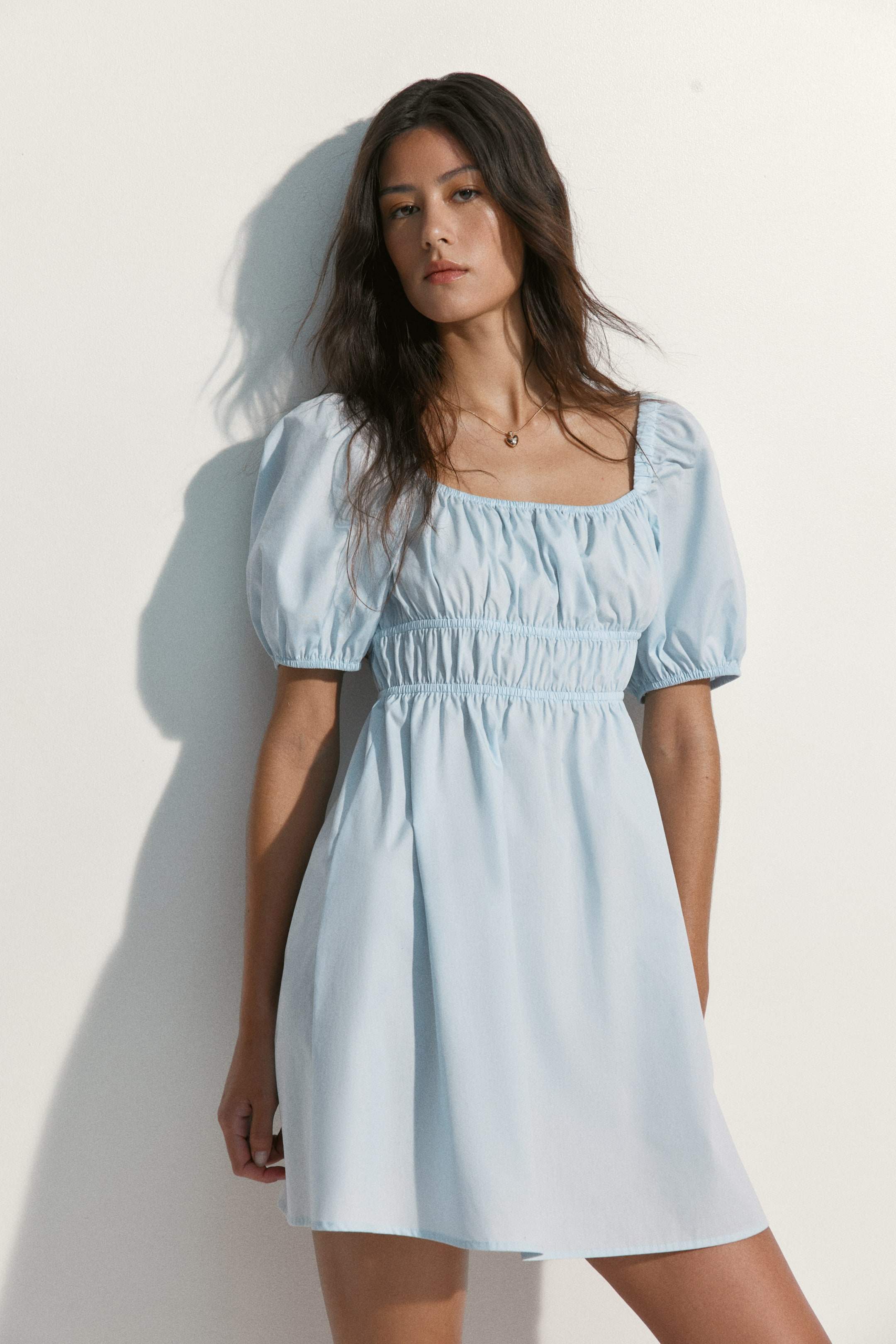 Ampliar la imagen: Un vestido mini de color azul claro presenta un escote cuadrado, mangas cortas abullonadas y un corpiño fruncido. Se ajusta en la cintura con pliegues y fluye hacia una falda acampanada.