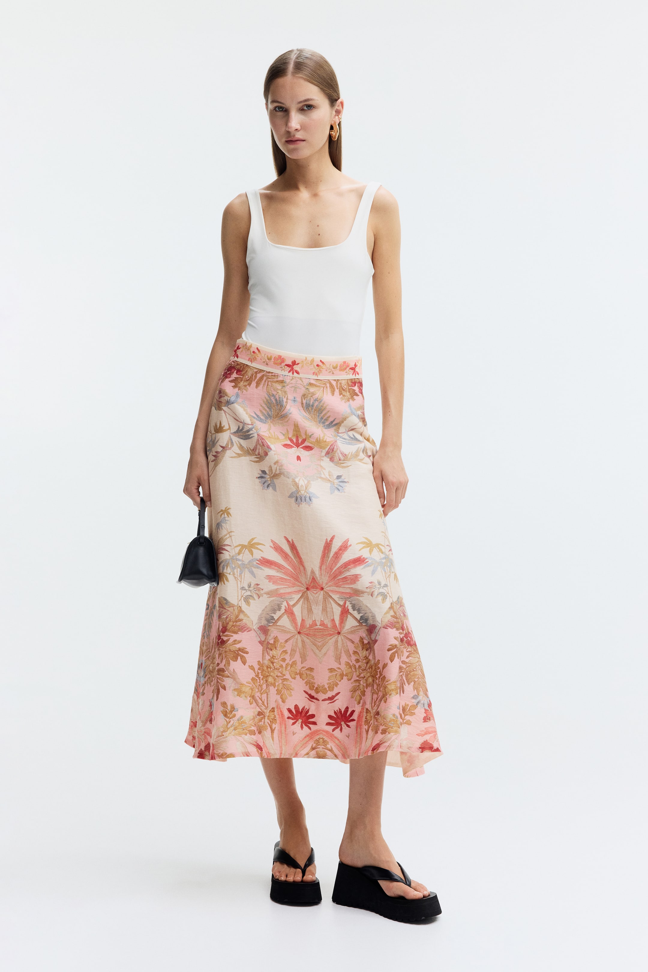 Visualizza immagine più grande: La modella indossa una gonna lunga con un tessuto croccante e una silhouette svasata, caratterizzata da una stampa botanica in rosa e beige. La gonna è abbinata a un top bianco con scollo quadrato e infradito con zeppa nere, mentre la modella tiene in mano una piccola borsa nera.