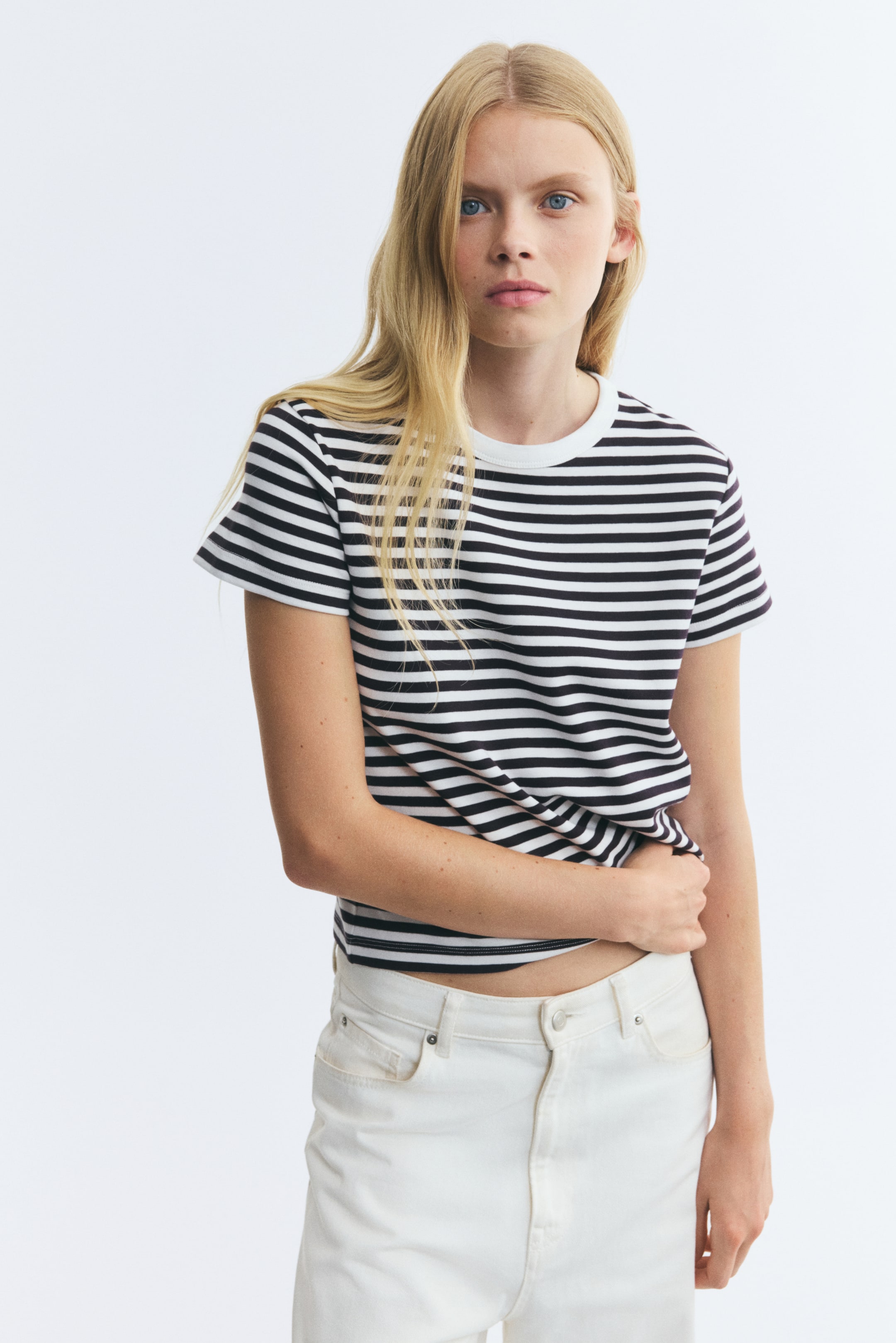 View larger image: A fitted, short-sleeved T-shirt featuring black and white horizontal stripes and a white ribbed crew neckline is being worn by the model. The model is styling the tee with white jeans and is resting one hand near the straight-cut hem.