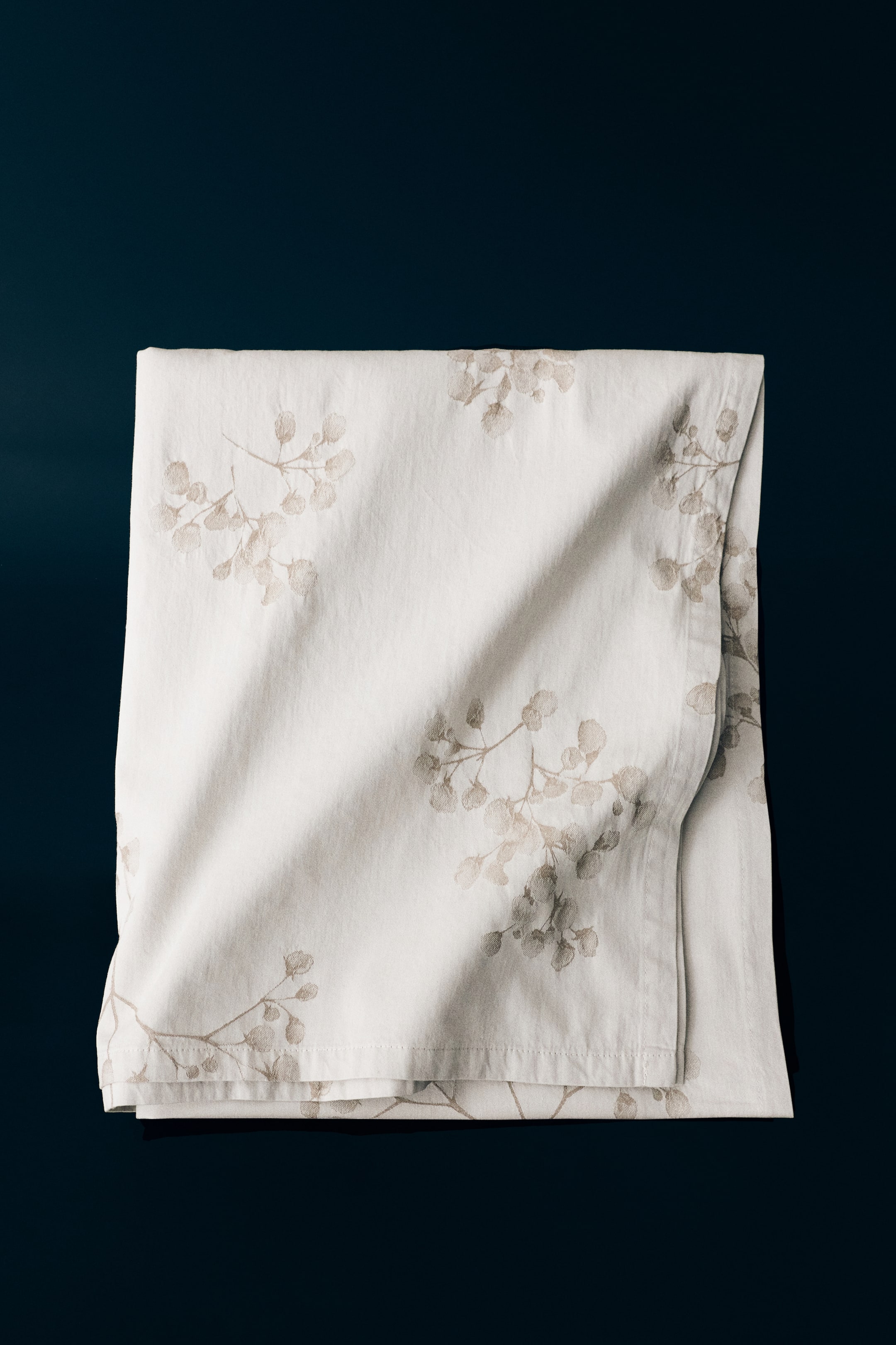 View larger image: A folded tablecloth in off-white cotton sateen displays a subtle jacquard-woven floral and leaf branch pattern in a muted beige tone. The tablecloth features soft, draped folds and clean-hemmed edges that highlight its smooth, high-thread-count texture.
