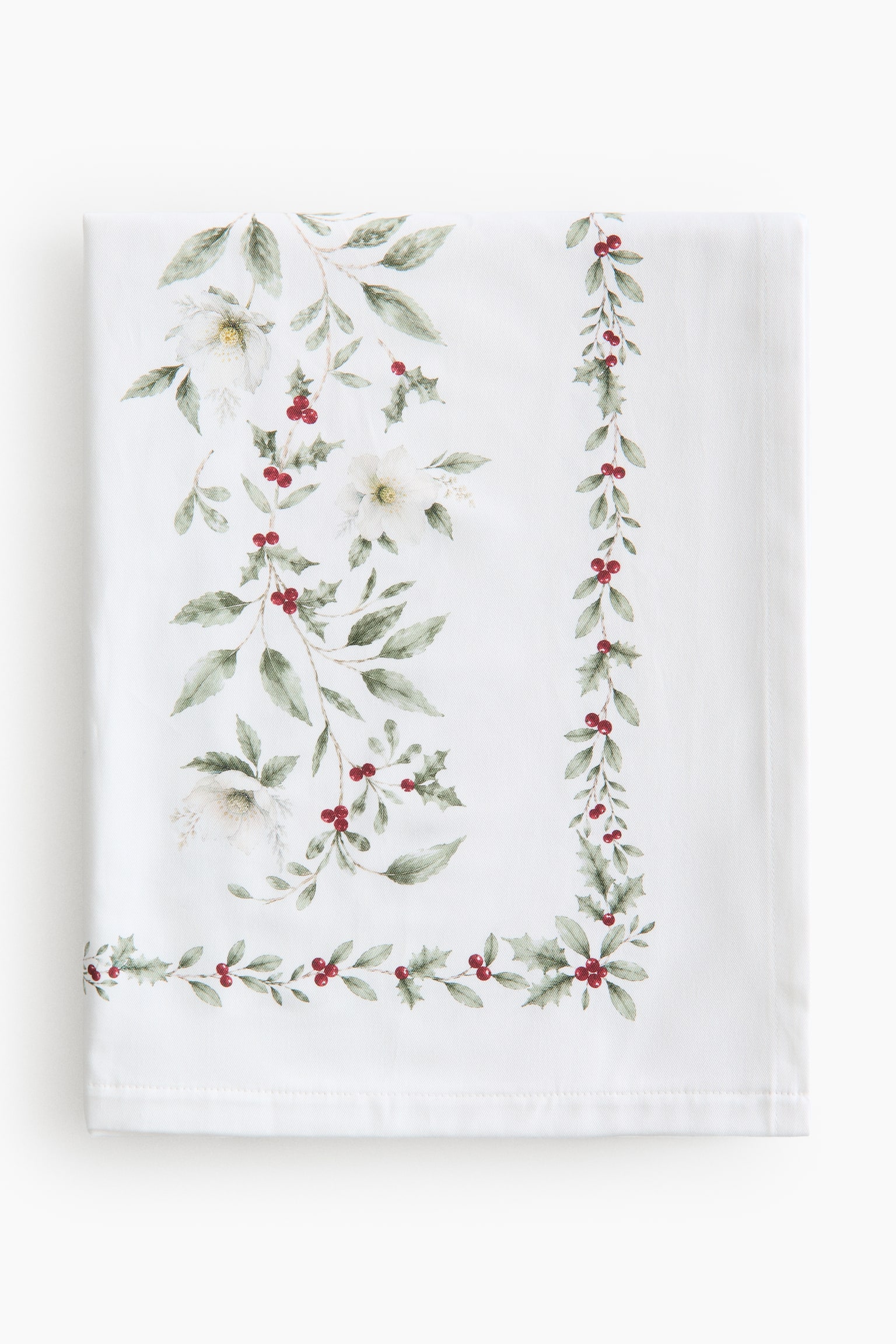 En vikt vit duk visar ett botaniskt mÃ¶nster med grÃ¶na blad, vita blommor och rÃ¶da bÃ¤r. Designen har en bÃ¥rd av jÃ¤rnek och rÃ¶da bÃ¤r lÃ¤ngs neder- och hÃ¶gerkanten, med en stÃ¶rre blomsterkvast av vita blommor och grÃ¶nt bladverk placerad till vÃ¤nster.