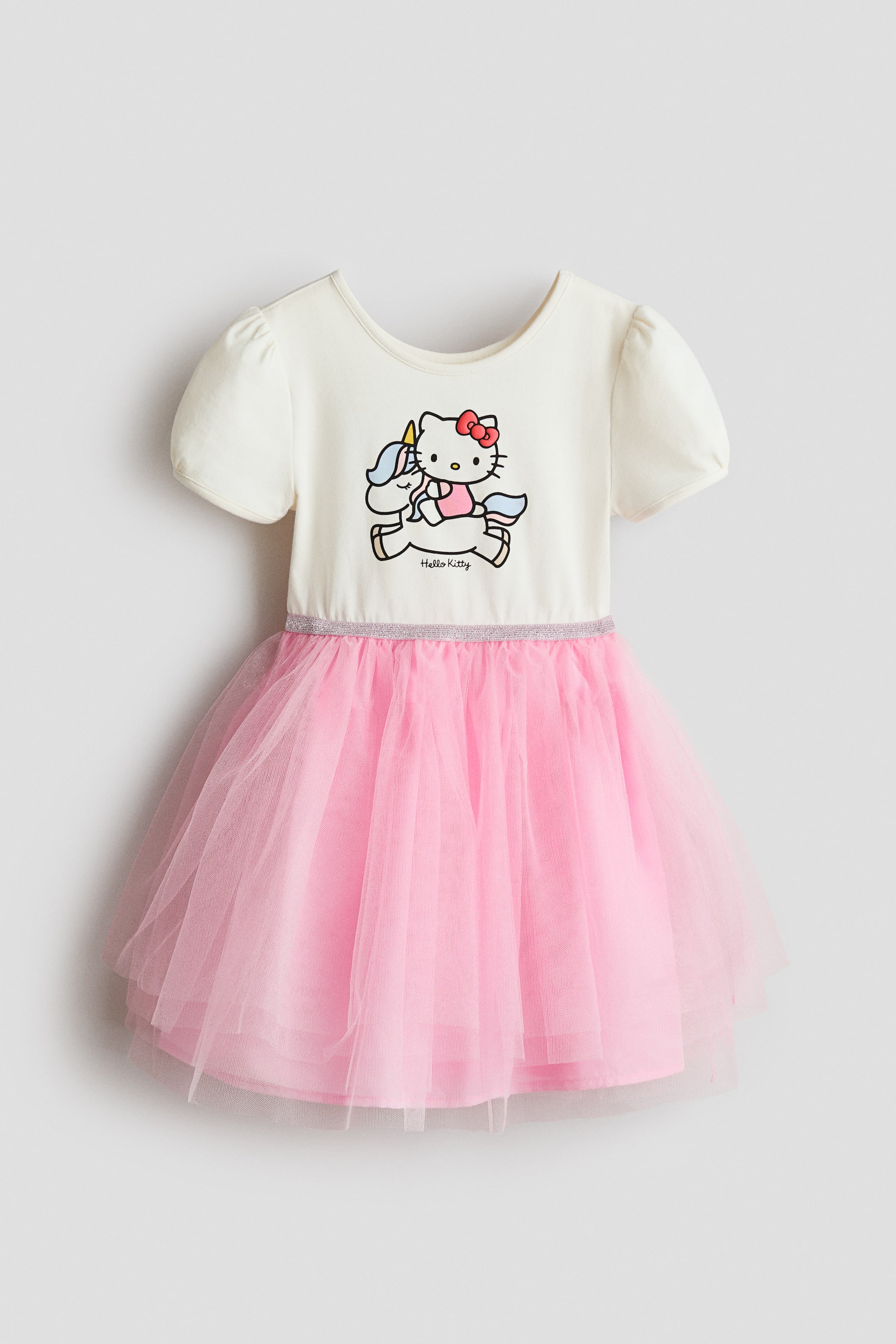 Voir une image plus grande: La robe présente un haut blanc cassé, des manches courtes bouffantes et un col rond, avec un motif Hello Kitty et licorne imprimé à l'avant. Une fine bande de paillettes argentées sépare le haut de la jupe en tulle rose à volants et froncée.