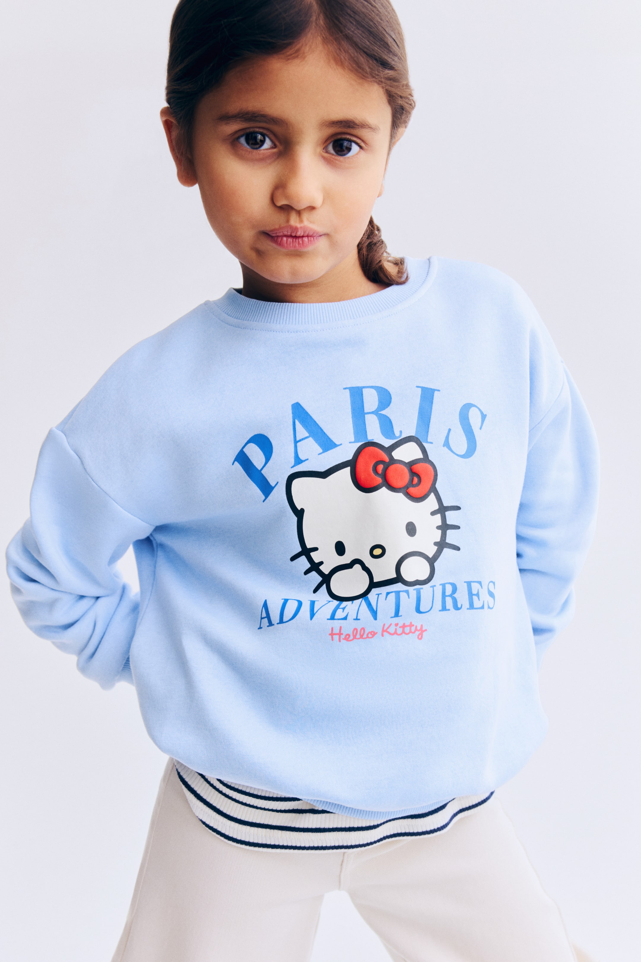 Ver imagen más grande: Sudadera azul claro de cuello redondo con un gran estampado gráfico en la parte delantera. El estampado muestra la palabra "PARIS" en letras azules arqueadas sobre la cabeza del personaje Hello Kitty con un lazo rojo, con la palabra "ADVENTURES" en letras azules y "Hello Kitty" en escritura rosa debajo. Diseñada con puños y bajo de canalé, con una prenda de rayas superpuesta por encima.