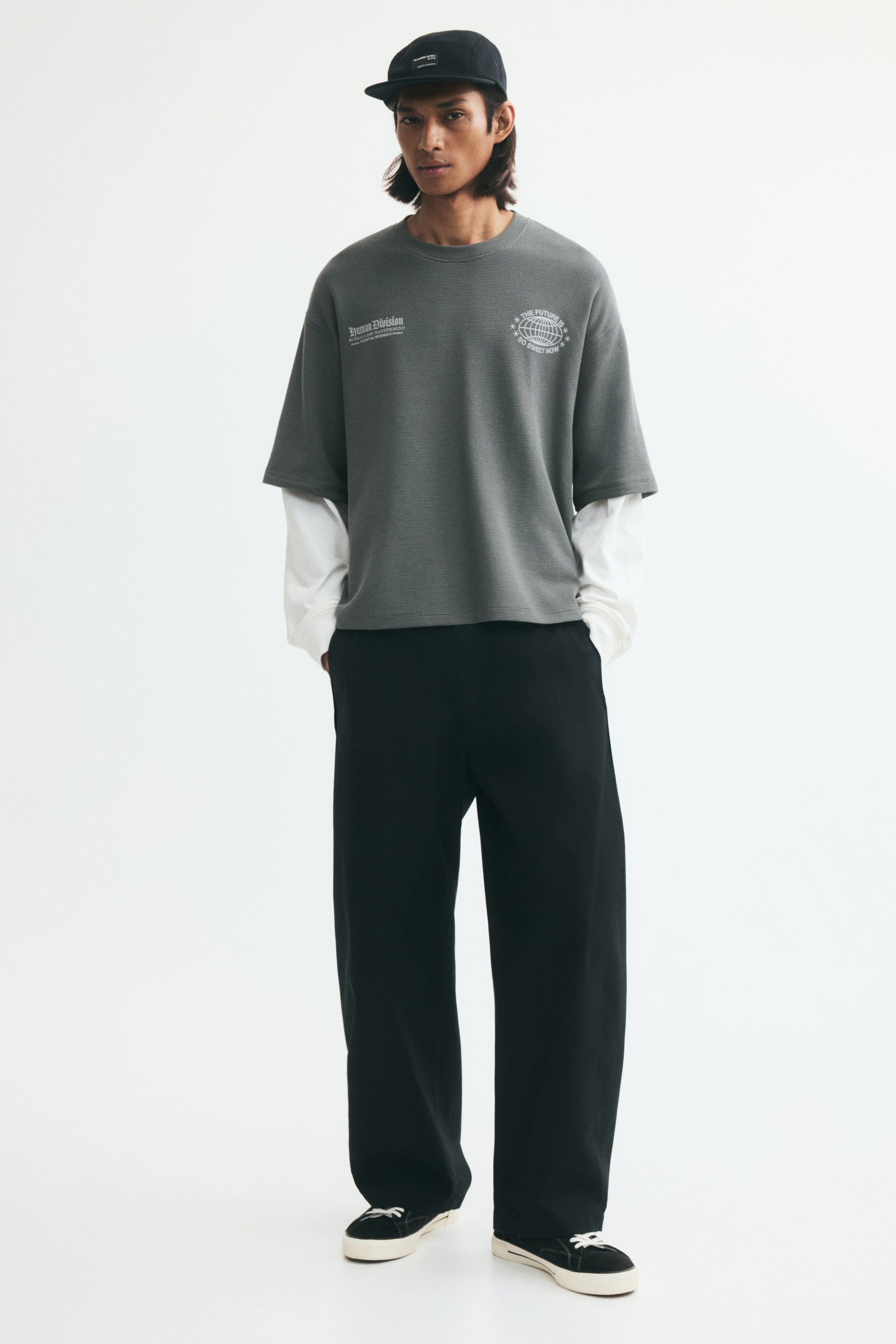 Agrandir l'image: Un t-shirt en coton gris texturé, oversize, avec un motif blanc sur la poitrine et de longues manches blanches intégrées en superposition est porté par le mannequin. Le t-shirt est assorti à un pantalon noir à jambes larges, des baskets noires et une casquette noire.