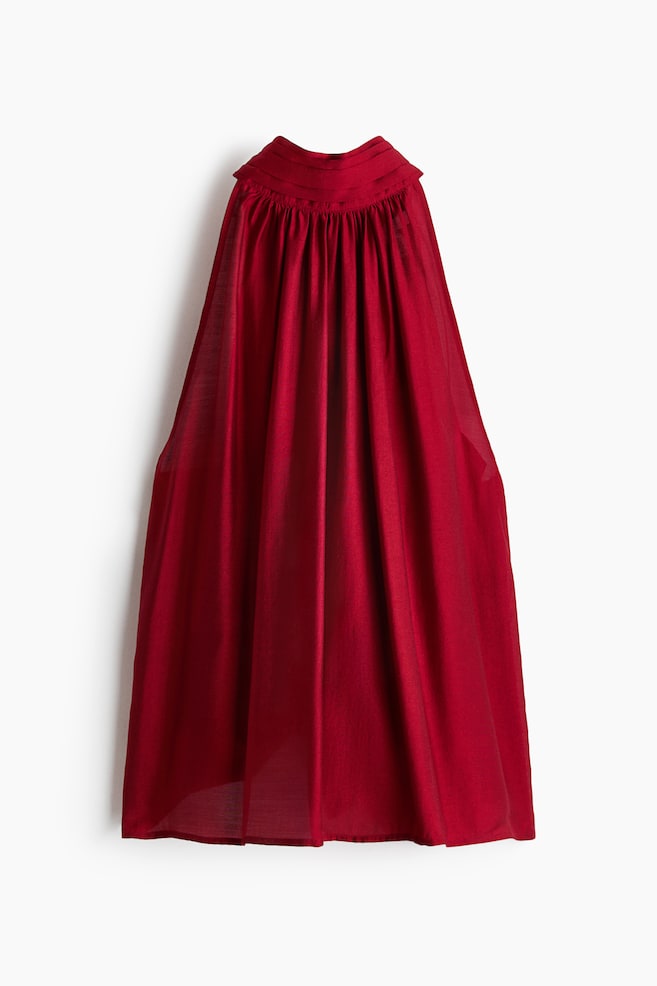 A sleeveless, A-line top in a deep red weave with a slight sheen is hanging facing the front. The design is showcasing a pleated stand collar with gathered fabric draping down to a straight-cut hem. - 1