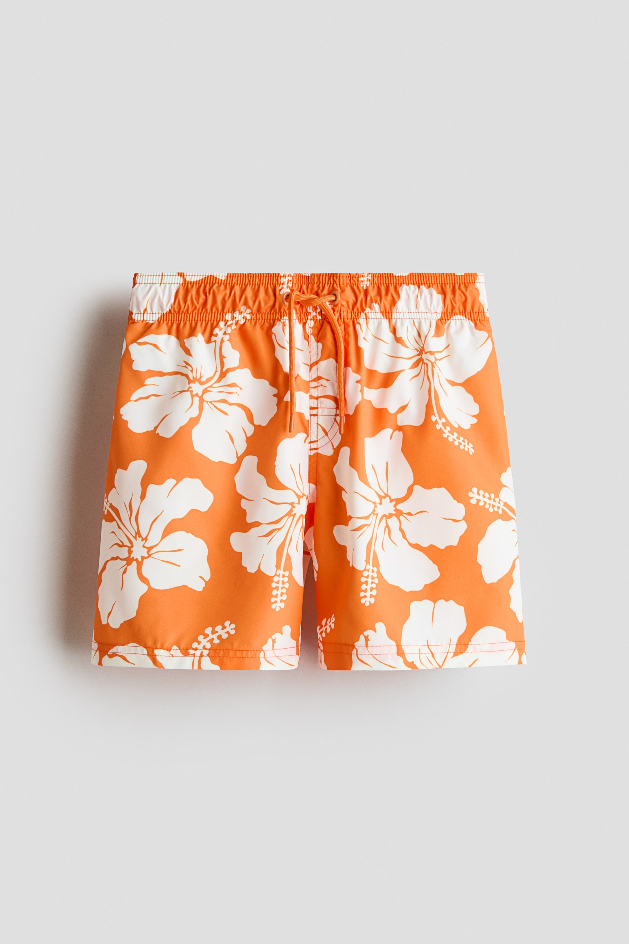Ver imagem maior: Os calções de banho são laranja com um estampado floral de hibisco branco, apresentando uma cintura elástica com um cordão branco atado num nó. São apresentados de frente.