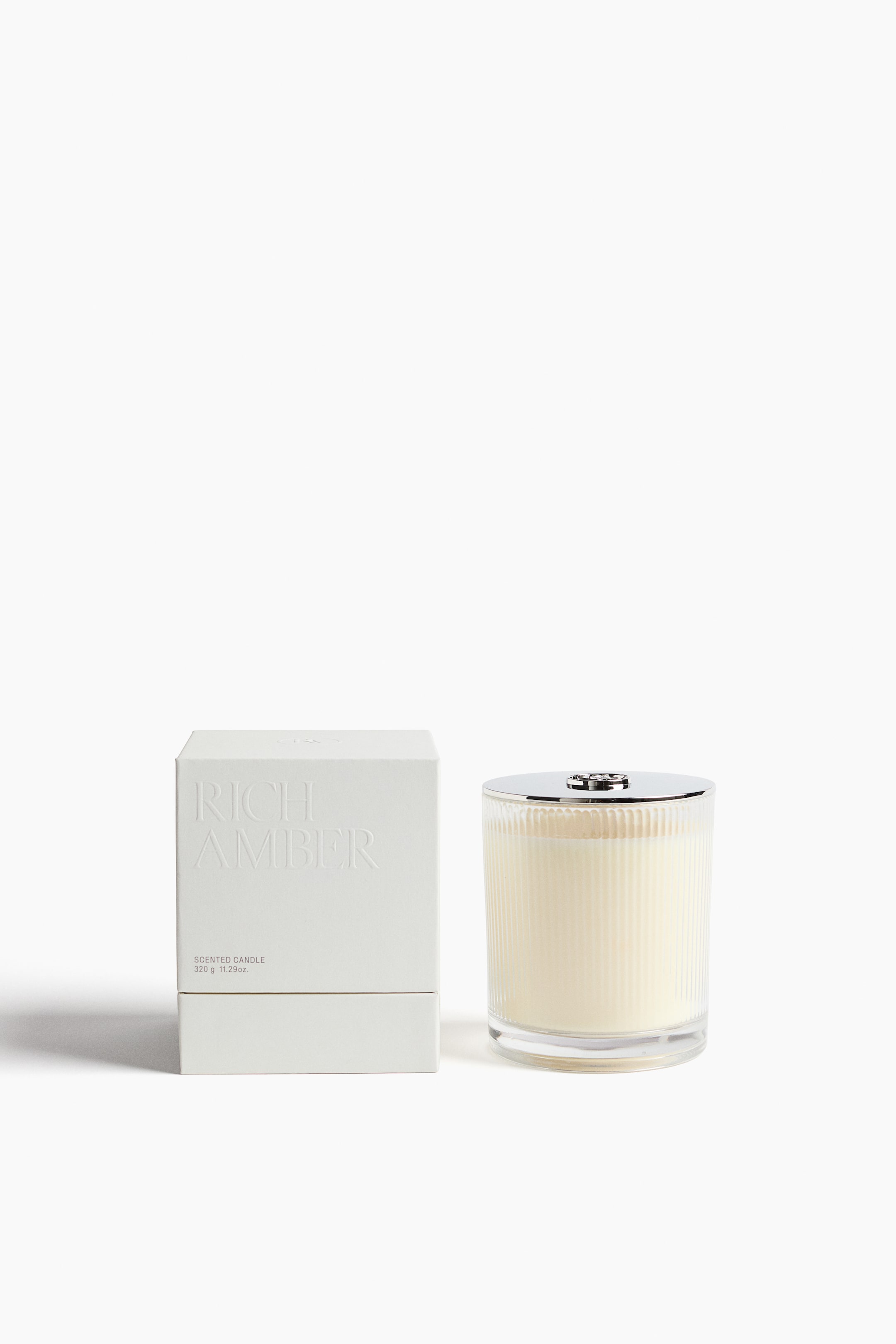 Agrandir l'image: Une vue de face montre une bougie parfumée placée à côté de son emballage carré blanc. La bougie présente un pot en verre nervuré verticalement, rempli de cire de couleur crème et surmonté d’un couvercle argenté poli, tandis que la boîte texturée affiche un lettrage en relief creusé indiquant « RICH AMBER ».