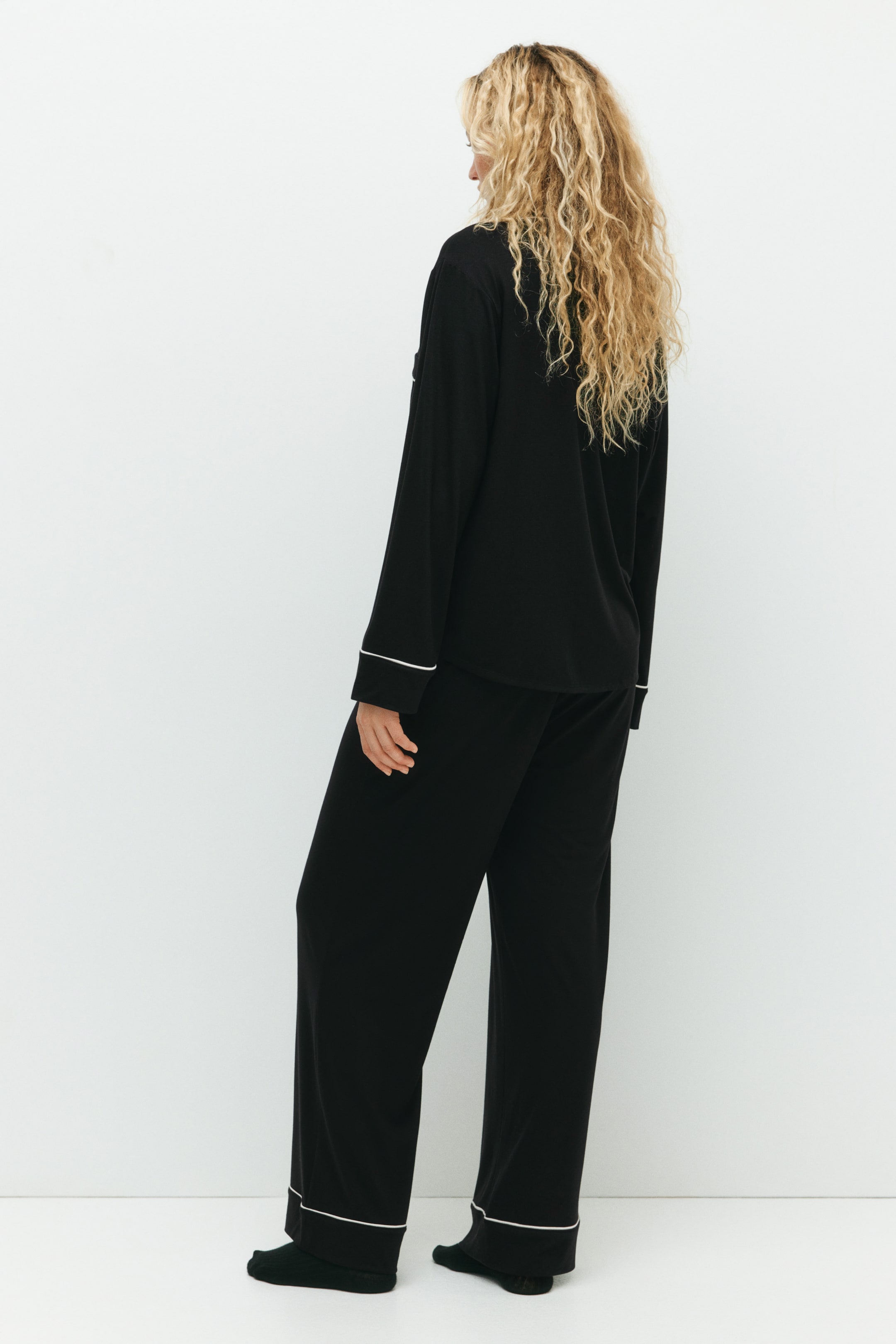 Größeres Bild ansehen: Ein schwarzes Pyjama-Set, gefertigt aus weichem Jersey, wird vom Model getragen. Das langärmelige Oberteil und die lange Hose verfügen über zarte weiße Paspeln an den Bündchen und Säumen.