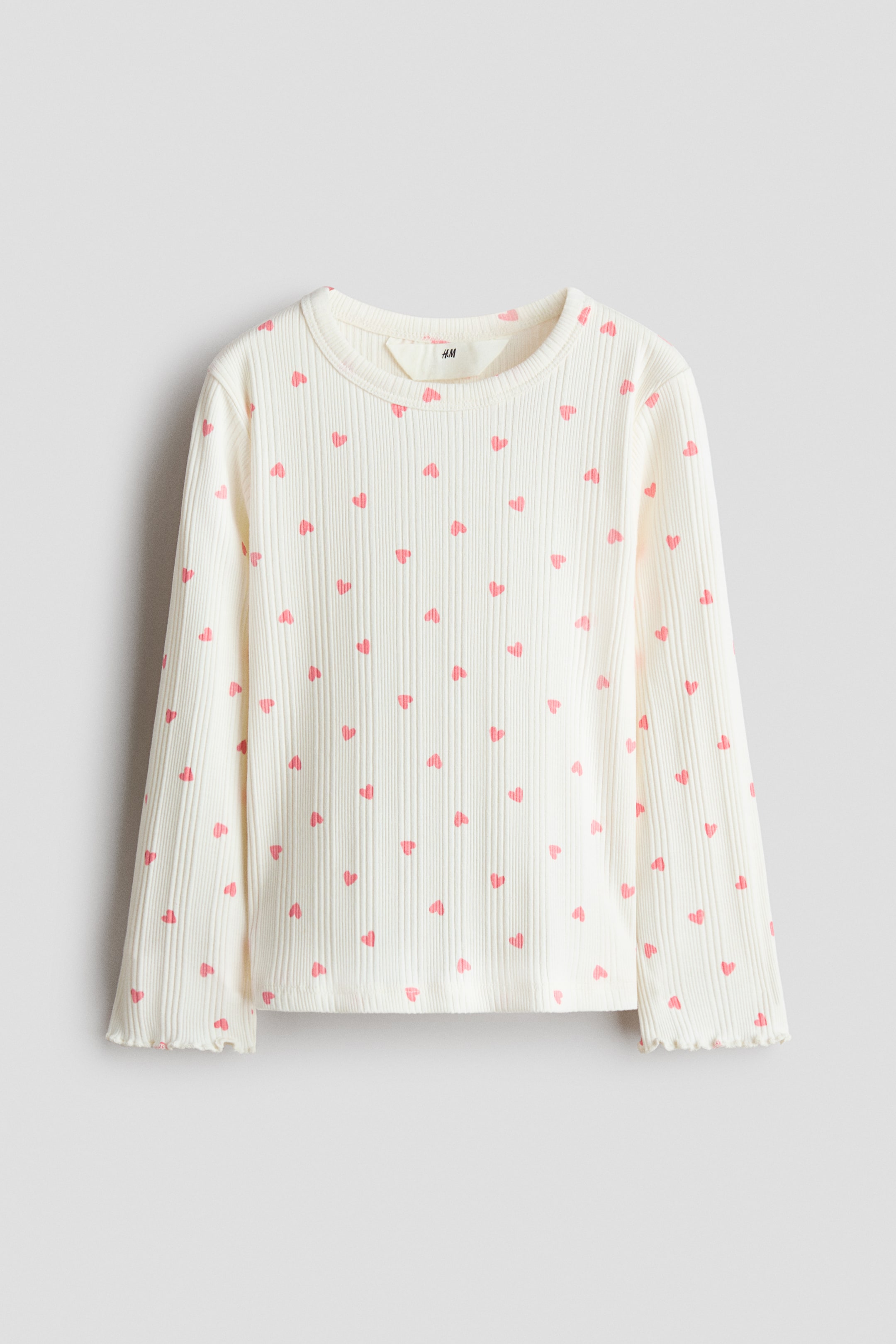 View larger image: A long-sleeved top is lying flat, featuring an off-white ribbed cotton jersey with an all-over pink heart print, a round neckline with a narrow trim, and overlocked edges at the cuffs.