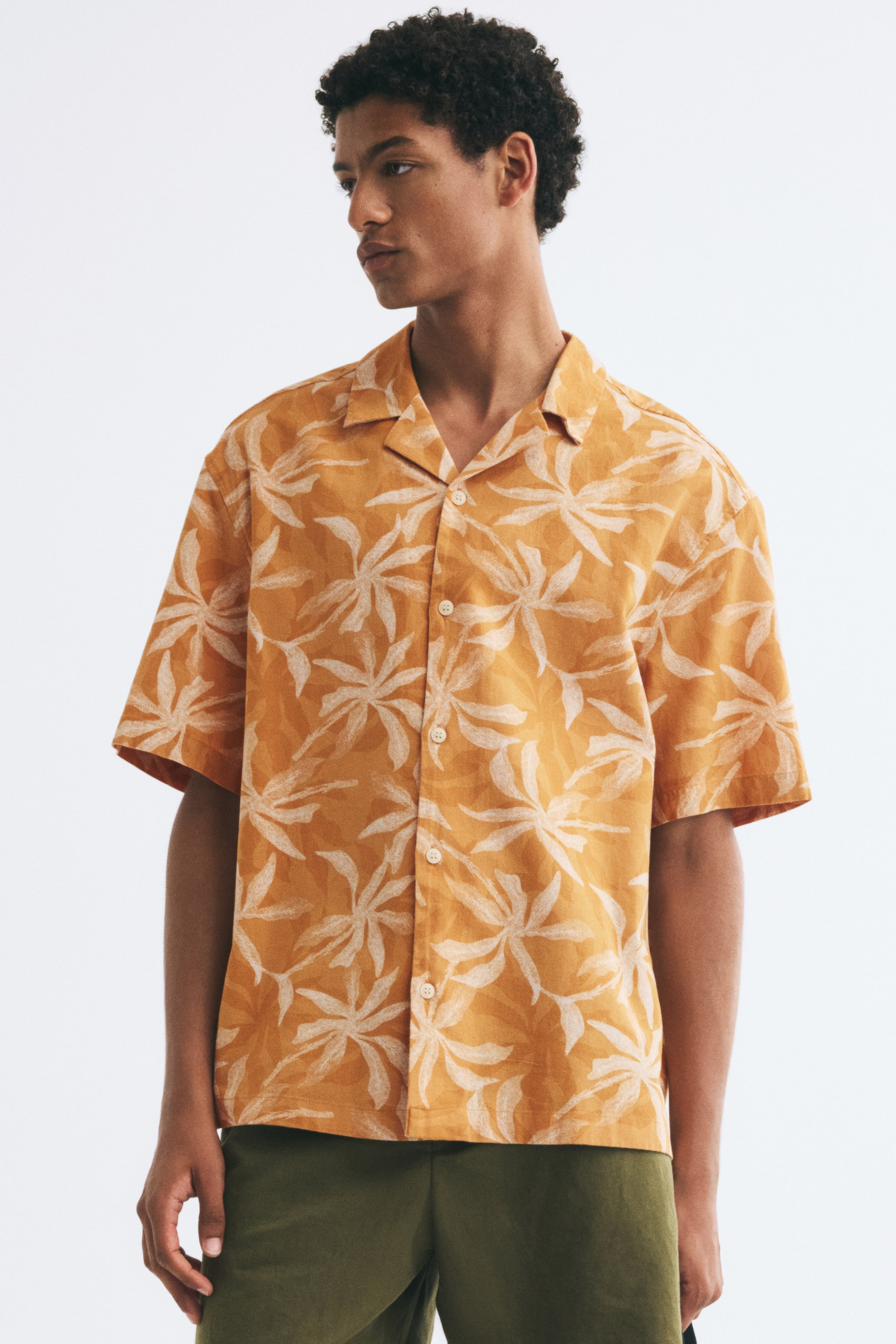 View larger image: The model is wearing a short-sleeve, relaxed-fit resort shirt in a mustard-yellow and white leaf pattern. The shirt features a resort collar and button-up front, and is paired with olive green trousers.