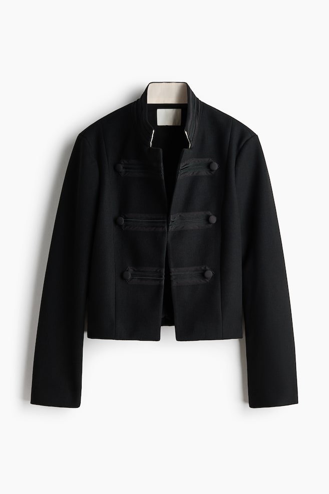 A black cropped jacket is hanging, facing forward, featuring a ribbed stand-up collar and an open front with three horizontal bands of black ribbon trim and round black buttons on each side. - 1