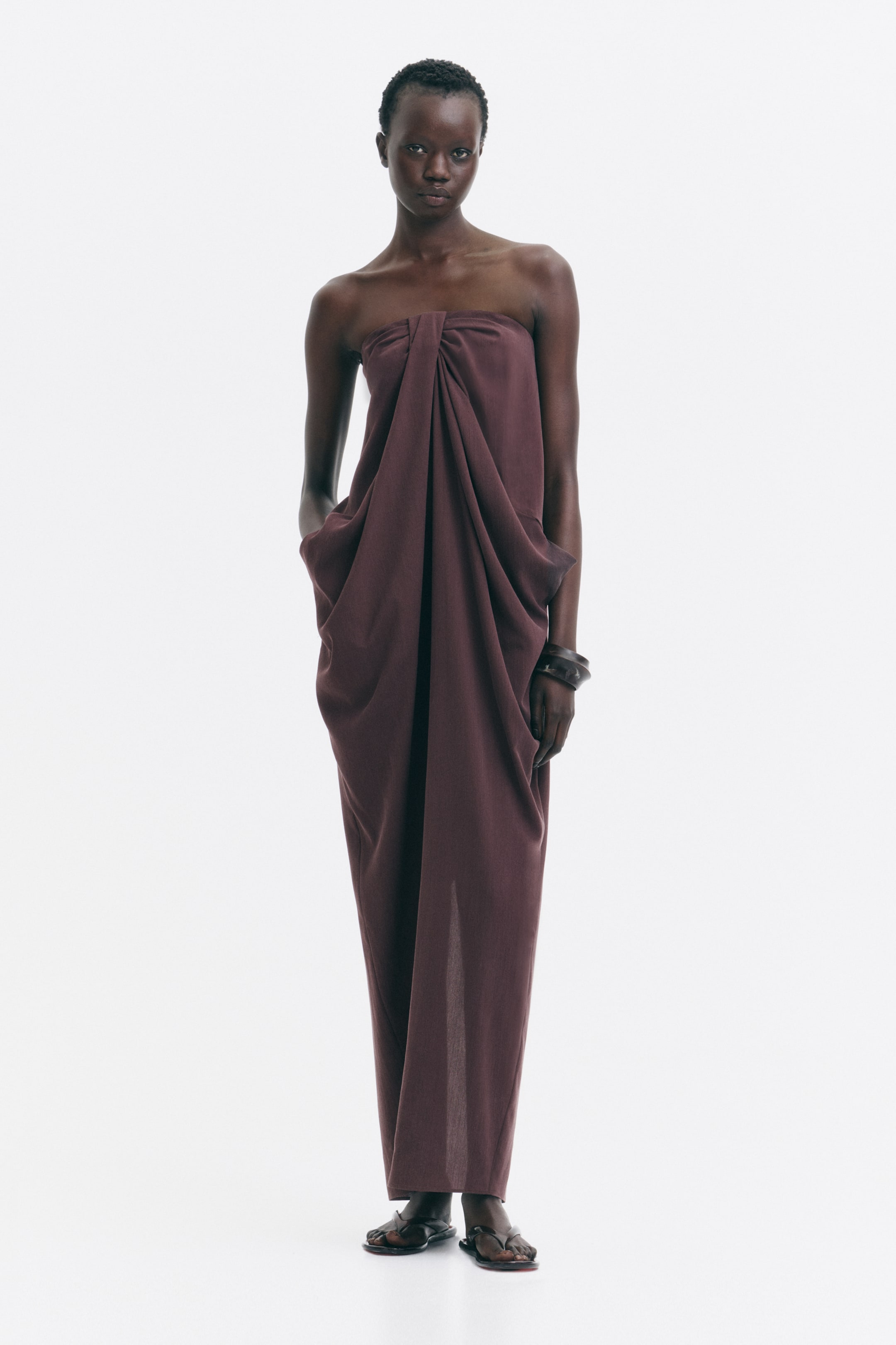 View larger image: The model is wearing a long, dark reddish-brown bandeau dress crafted from a lyocell-blend weave with a subtle sheen. It showcases a striking knot detail at the front, creating a voluminous draped effect. The dress also features discreet side pockets where the model is placing her hands.
