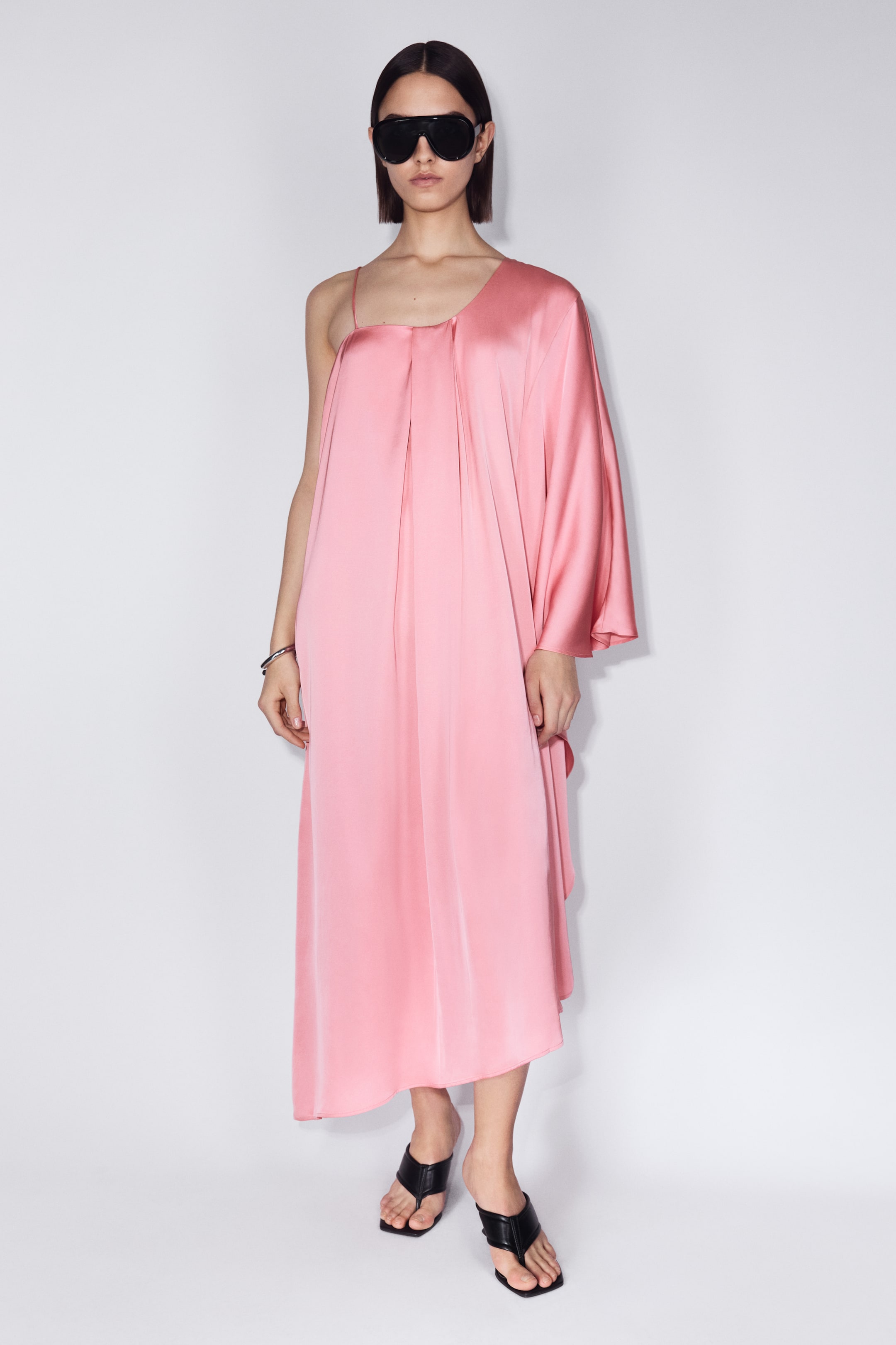 Ampliar la imagen: Un vestido midi rosa de un solo hombro presenta una silueta drapeada con una manga larga y un tirante delgado en el hombro opuesto. Está combinado con sandalias negras de dedo con punta cuadrada y gafas de sol negras de escudo sobredimensionado.