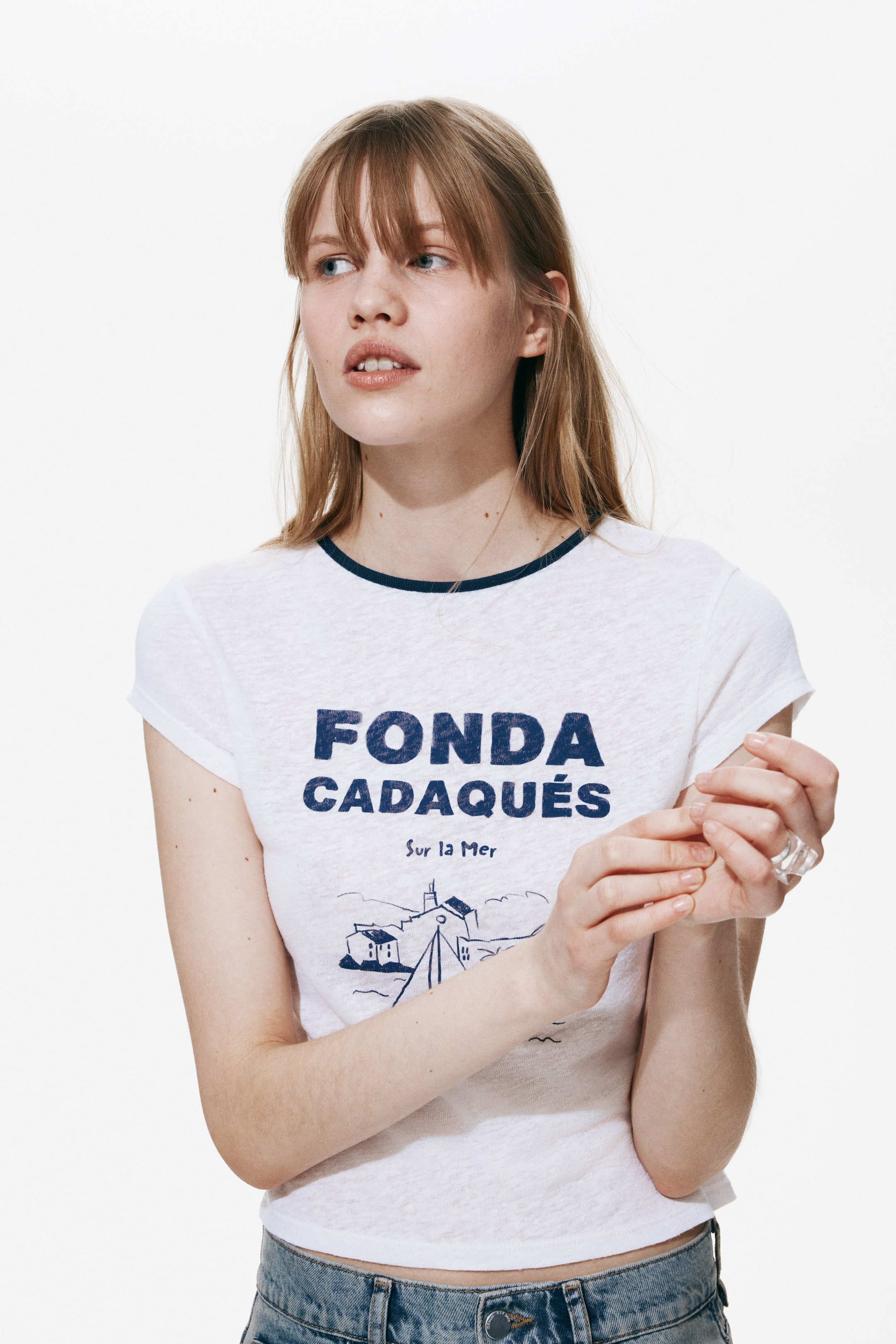 View larger image: The white t-shirt features a dark blue trimmed round neckline and short sleeves. It displays a dark blue graphic print on the front, reading 'FONDA CADAQUÉS Sur la Mer' with a coastal illustration, and its hem rests just above blue denim jeans.