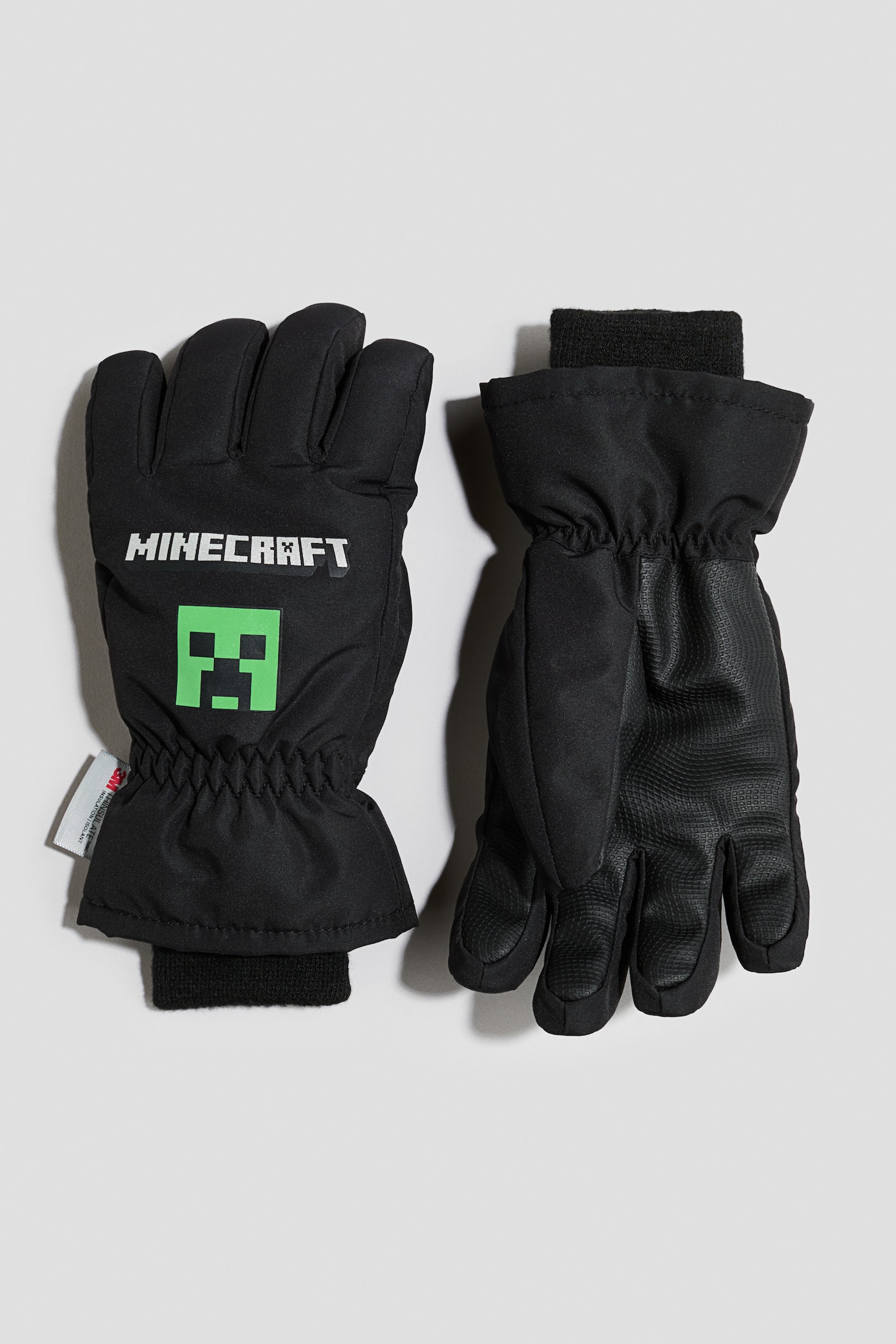 Une paire de gants dâhiver noirs est prÃ©sentÃ©e Ã  plat, le gant de gauche montrant le design de lâavant avec un logo blanc Â« MINECRAFT Â» et un graphique vert de Creeper. Le gant de droite est retournÃ© pour montrer lâadhÃ©rence texturÃ©e sur la paume et les doigts, les deux gants prÃ©sentant des poignets Ã©lastiquÃ©s et des poignets intÃ©rieurs cÃ´telÃ©s noirs.