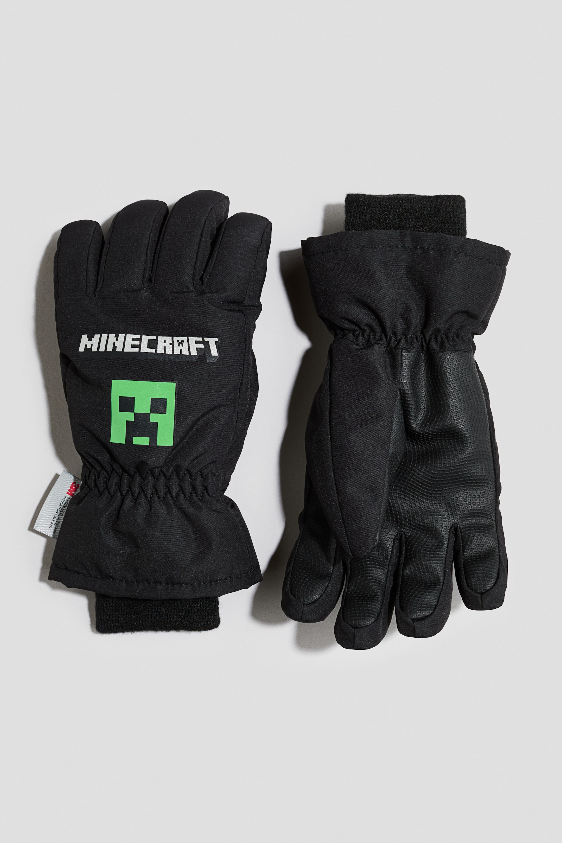 Voir une image plus grande: Une paire de gants d’hiver noirs est présentée à plat, le gant de gauche montrant le design de l’avant avec un logo blanc « MINECRAFT » et un graphique vert de Creeper. Le gant de droite est retourné pour montrer l’adhérence texturée sur la paume et les doigts, les deux gants présentant des poignets élastiqués et des poignets intérieurs côtelés noirs.