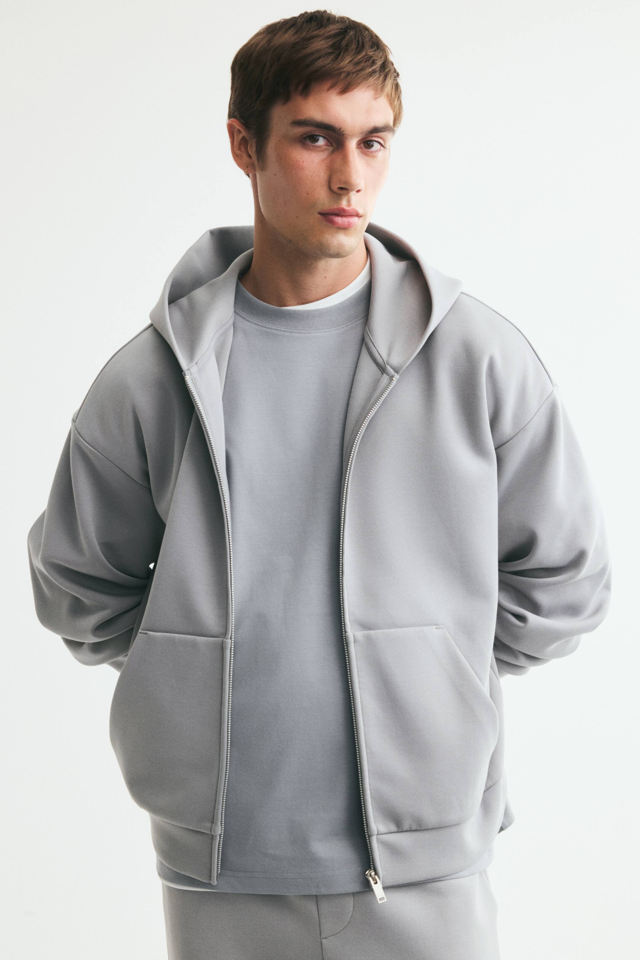 View larger image: A light gray zip-up hoodie in interlock jersey is being worn by the model. This oversized hoodie features a lined hood, a partially unzipped front, dropped shoulders, long sleeves, and front pockets where the model's hands are resting, and is layered over a matching crewneck top.