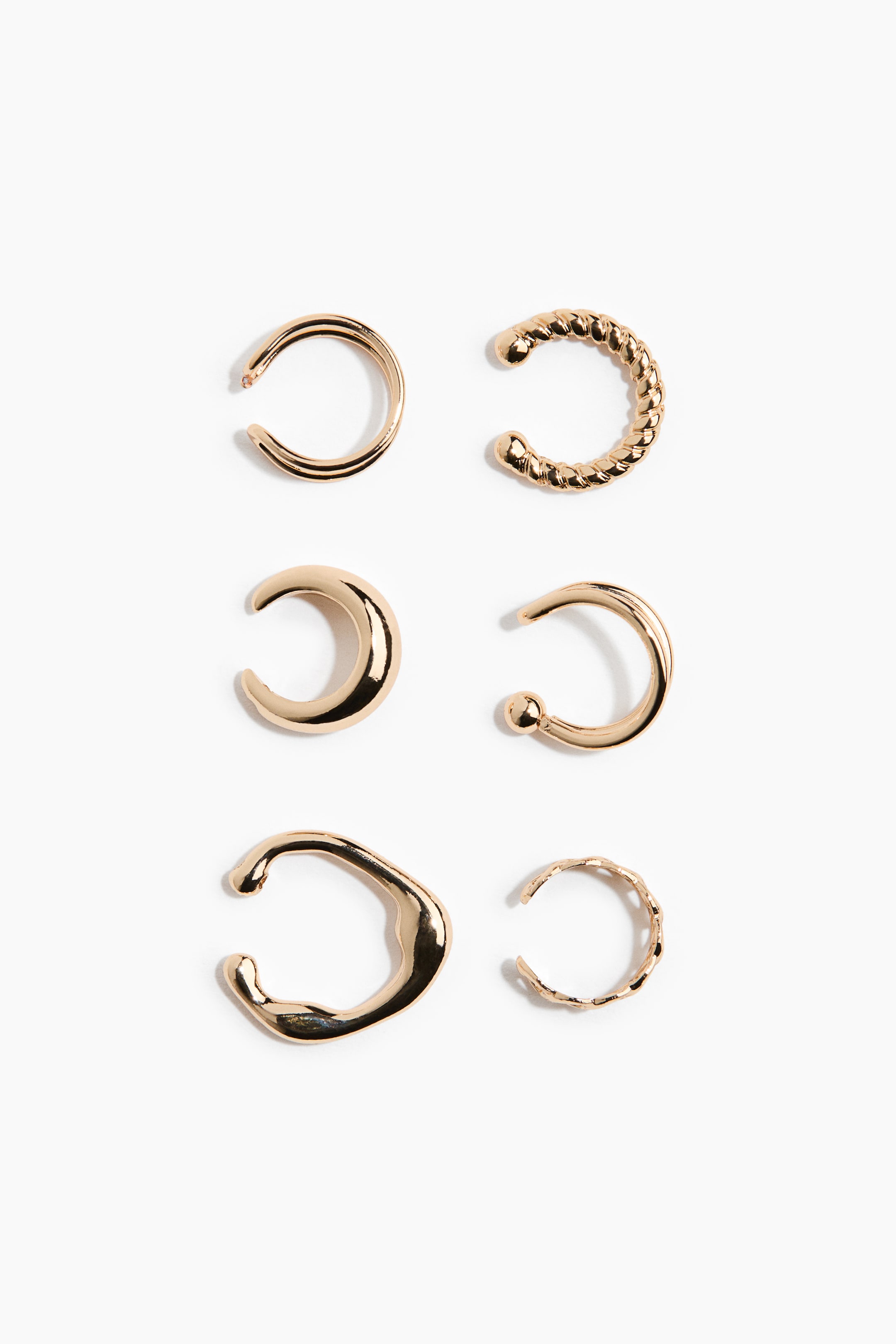 View larger image: Gold-toned metal ear cuffs in six distinct designs are displayed front-facing, arranged in two vertical columns. The collection features a variety of styles, including a classic smooth hoop, a twisted rope texture, a chunky crescent shape, a double-banded cuff with a beaded end, and two organic, wavy-textured bands.