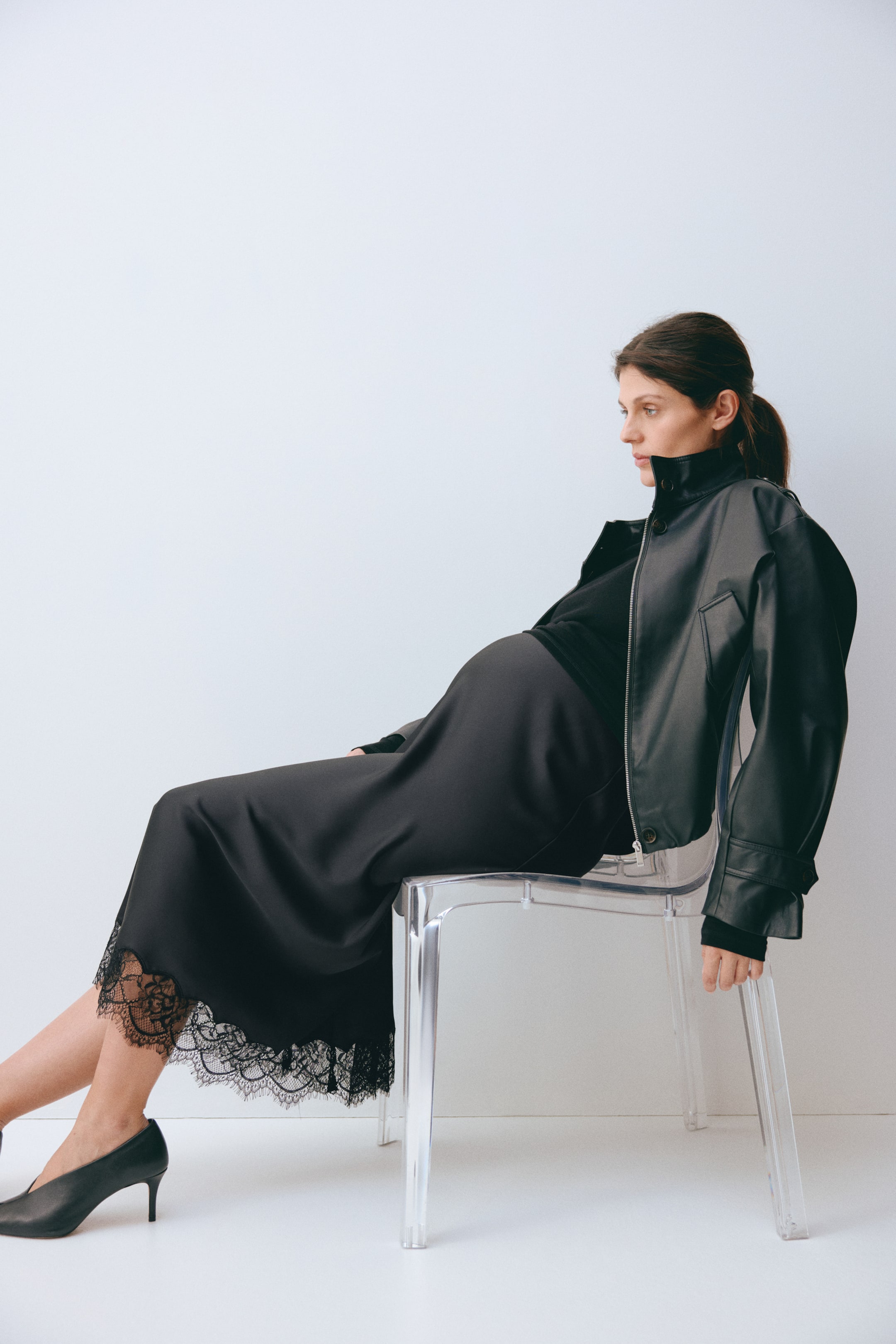 Ampliar la imagen: Se presenta un conjunto negro que incluye una chaqueta bomber de cuero sintético, un top de cuello alto, una falda midi de satén y tacones de punta afilada. La chaqueta destaca por su cuello alto, cierre frontal con cremallera y mangas voluminosas con detalles en los puños, mientras que la falda exhibe un amplio ribete de encaje negro a lo largo de su dobladillo desde un ángulo lateral.