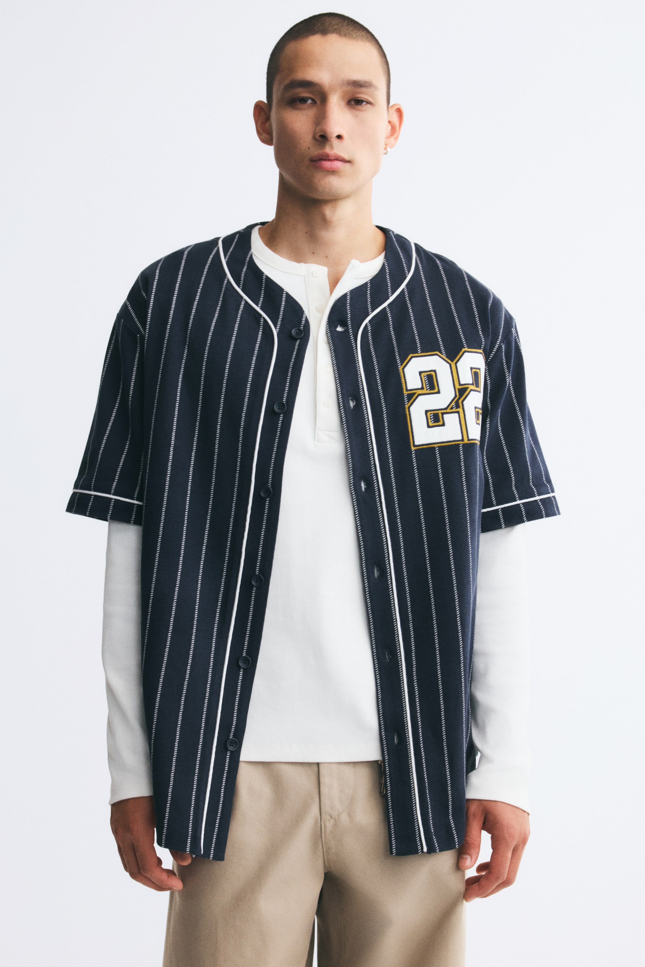 Ver imagen más grande: La modelo lleva abierta una camiseta de béisbol azul marino a rayas finas, que muestra ribetes blancos en el escote, la tapeta y los puños, y el número "22" impreso en el pecho. La camiseta de manga corta se lleva superpuesta sobre una prenda blanca de manga larga y se combina con un pantalón beige.