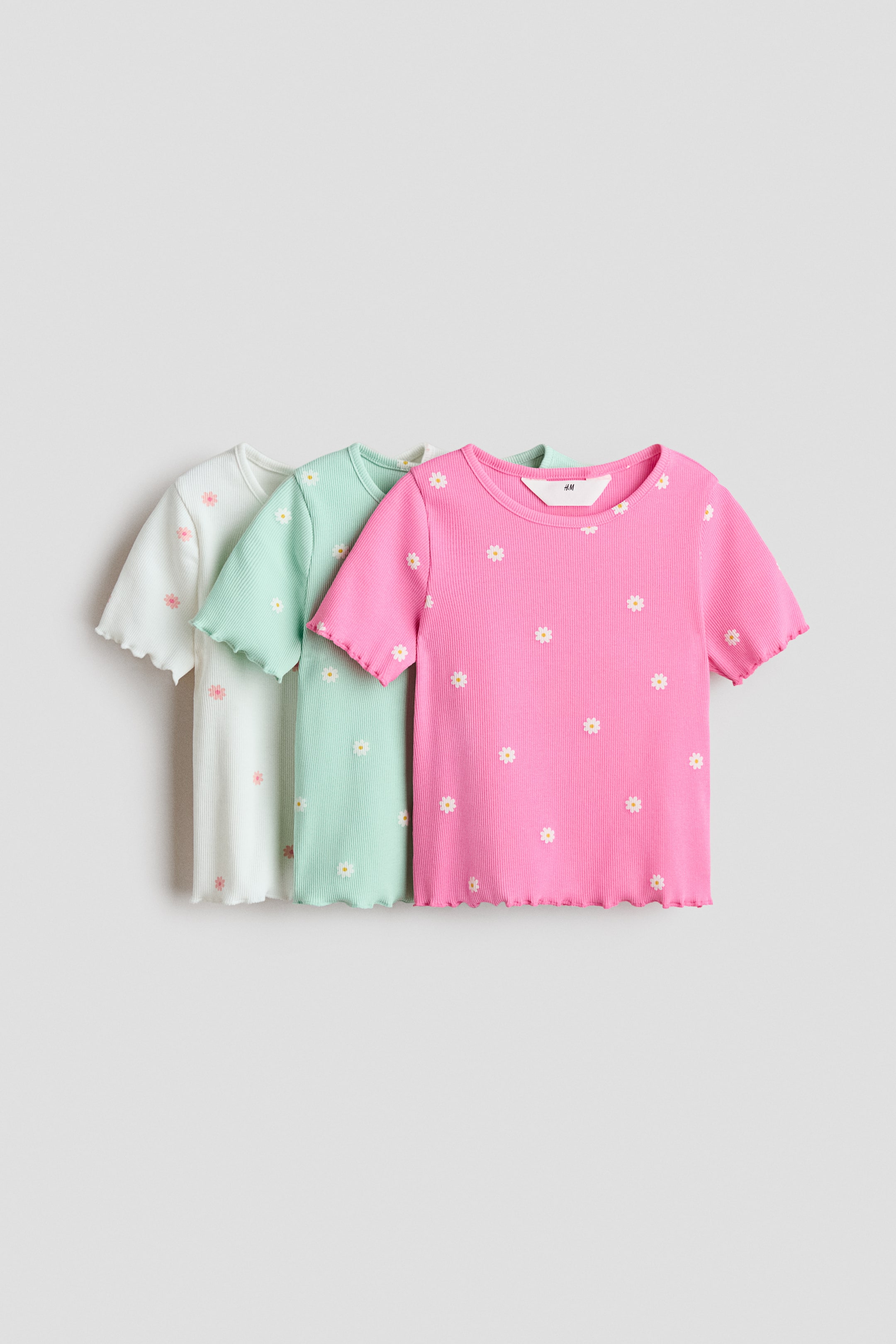 View larger image: Three ribbed t-shirts are displayed from a front angle, with each t-shirt slightly overlapping the one behind it. Each t-shirt features short sleeves, a round neckline, and scalloped hems on the sleeves and bottom. The leftmost white t-shirt showcases pink and yellow daisy patterns, the middle mint green t-shirt showcases white and yellow daisy patterns, and the rightmost hot pink t-shirt showcases white and yellow daisy patterns.