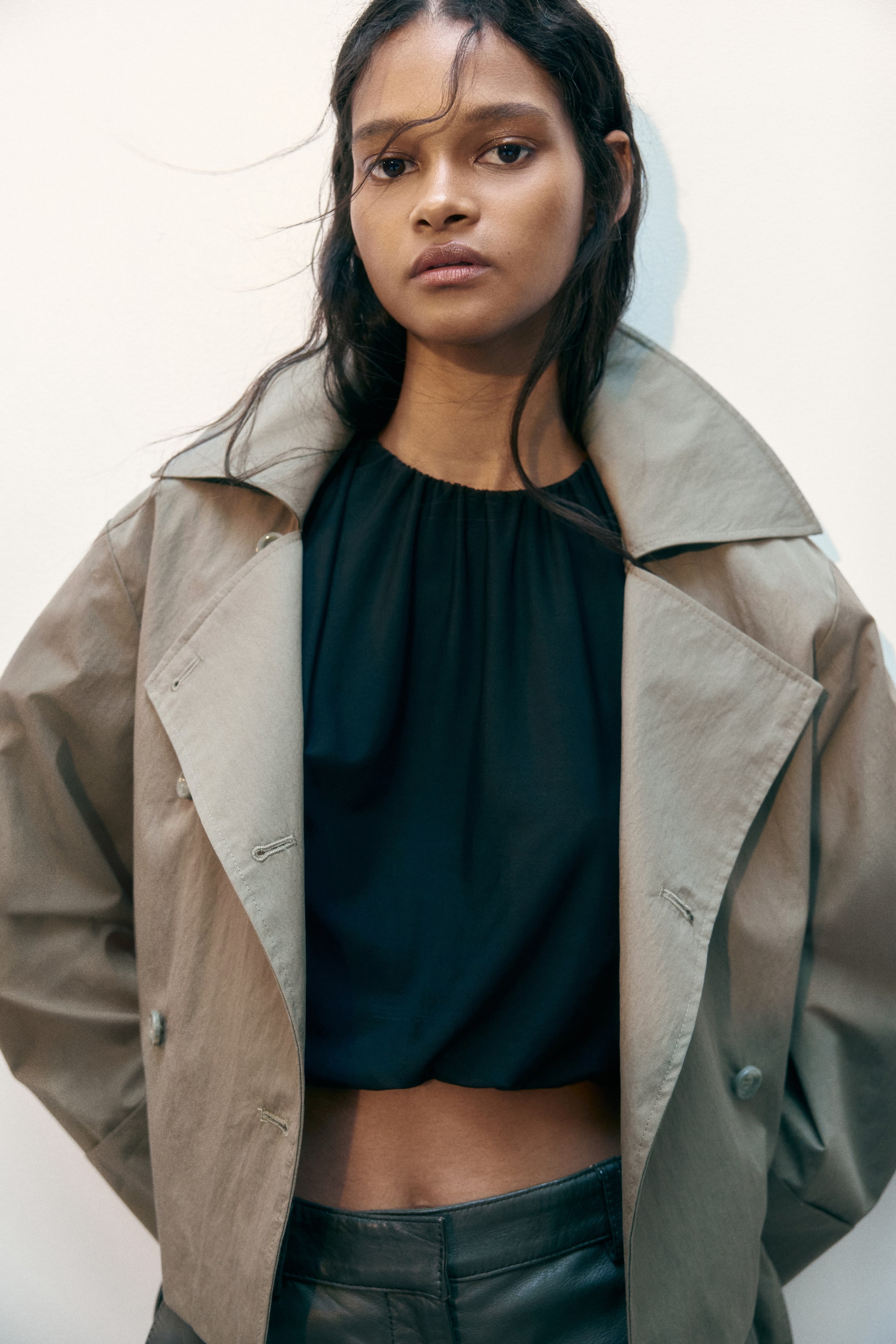 Grotere afbeelding bekijken: Een korte, lichtgekleurde double-breasted twill jas met een brede kraag, knopen aan de voorkant en verlaagde schouders wordt door het model open gedragen, waardoor een donkere top eronder zichtbaar is.