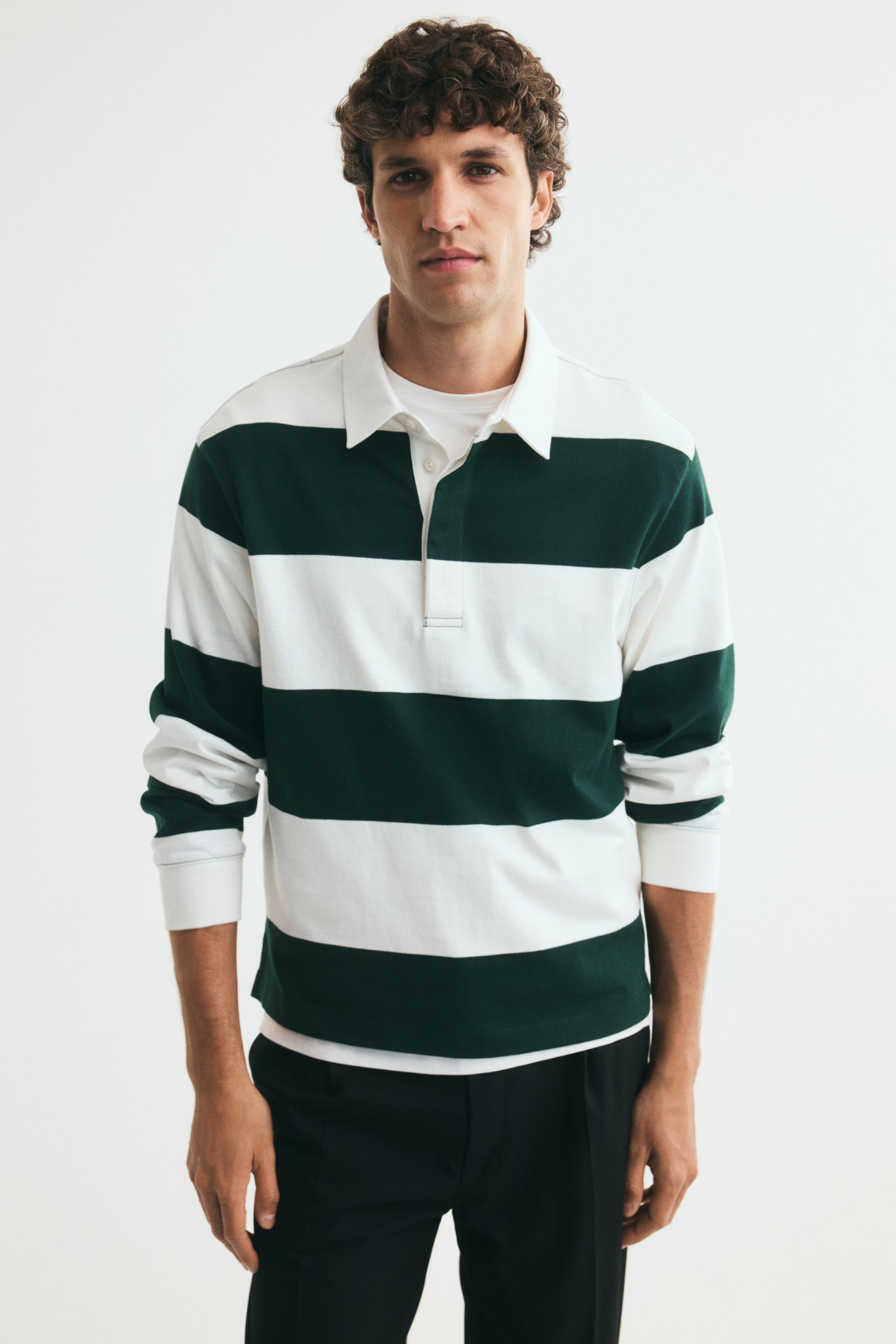 Voir une image plus grande: Le mannequin porte un polo de rugby rayé vert et blanc, doté d’un col rabattu blanc, d’une patte de boutonnage partiellement ouverte, de manches tombantes et de poignets côtelés. La chemise est superposée à un t-shirt blanc à col rond et agencée avec un pantalon noir.