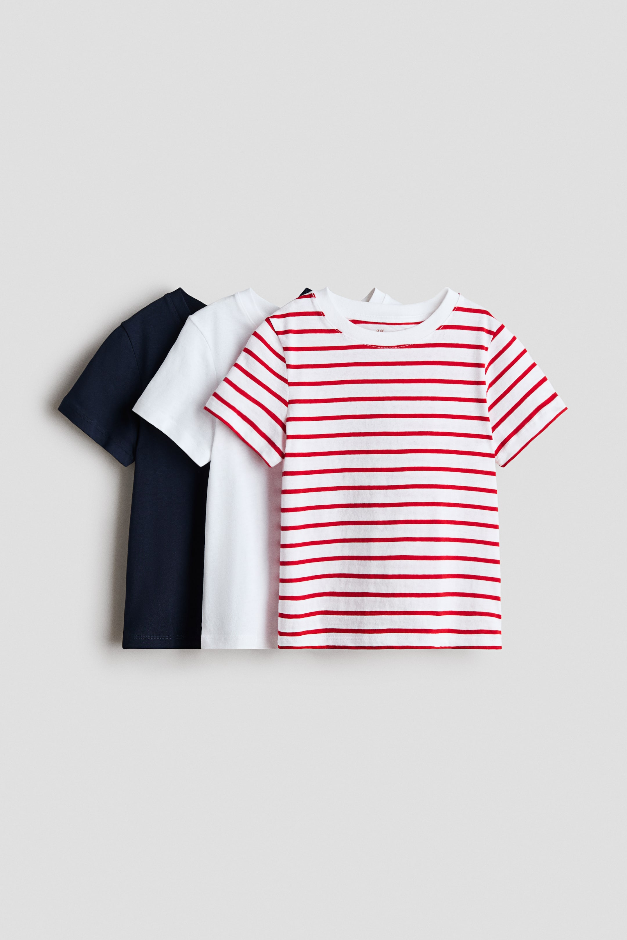 Visualizza l'immagine ingrandita: Tre t-shirt sono presentate da un'angolazione frontale, realizzate in morbido jersey di cotone e caratterizzate da un girocollo con bordo a costine. Sono disponibili con maniche corte, in blu navy, bianco semplice e bianco con righe orizzontali rosse.