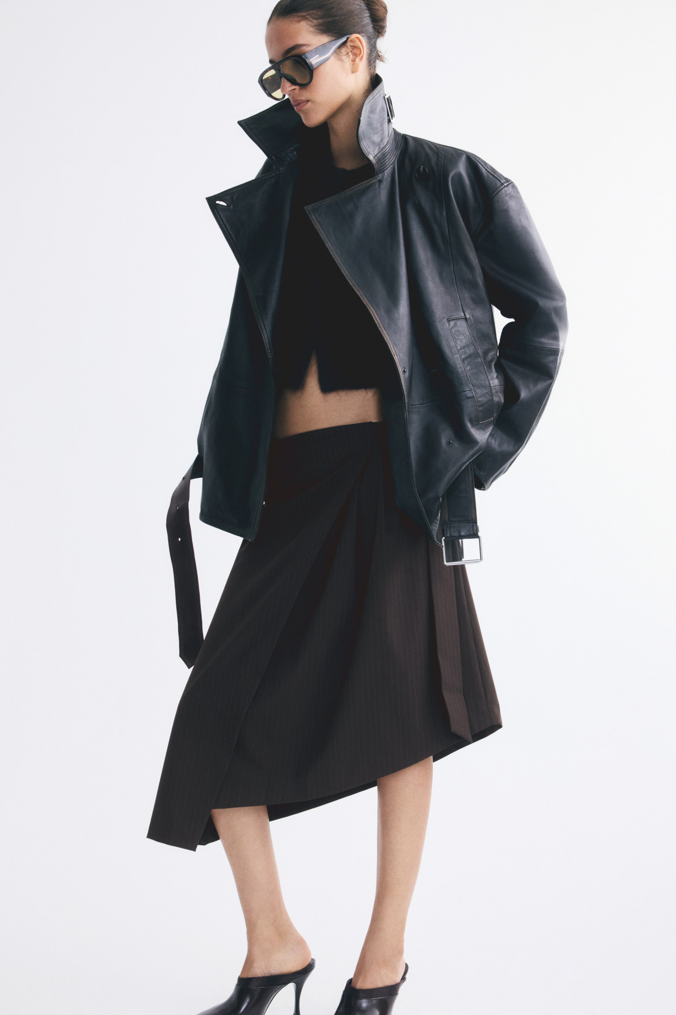 View larger image: A calf-length wrap skirt in dark brown woven fabric is worn by the model, featuring front pleats, an adjustable waist belt, and an asymmetrical hem. The skirt is styled with a black crop top, an oversized black leather jacket, and black heeled mules.