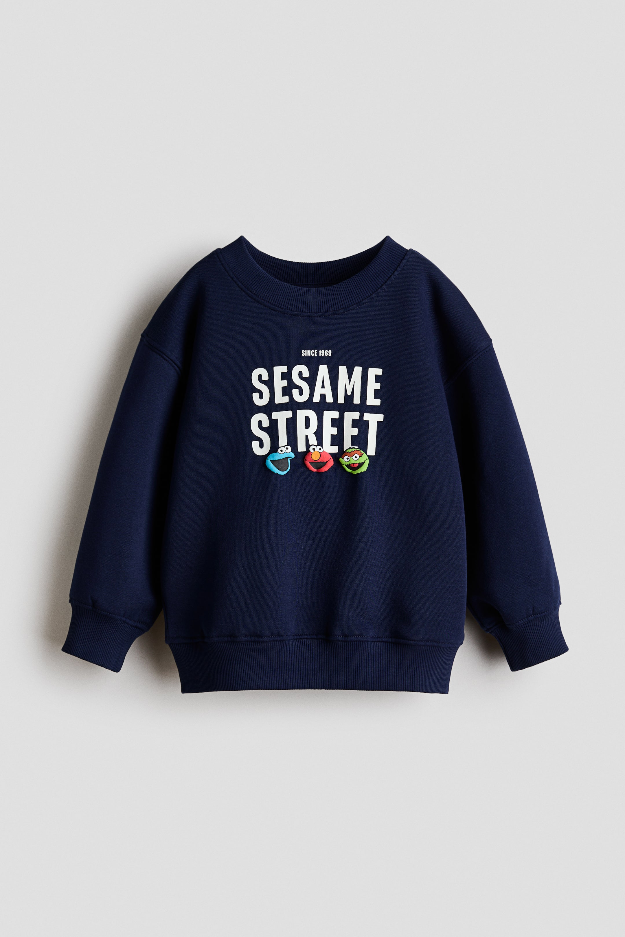 Grotere afbeelding bekijken: Een marineblauwe sweatshirt toont zijn voorkant, met een witte "SINCE 1969 SESAME STREET" print en drie kleurrijke karakterhoofden eronder. Het heeft een ronde hals met ribboord, verlaagde schouders, lange mouwen met geribde manchetten en een geribde zoom.