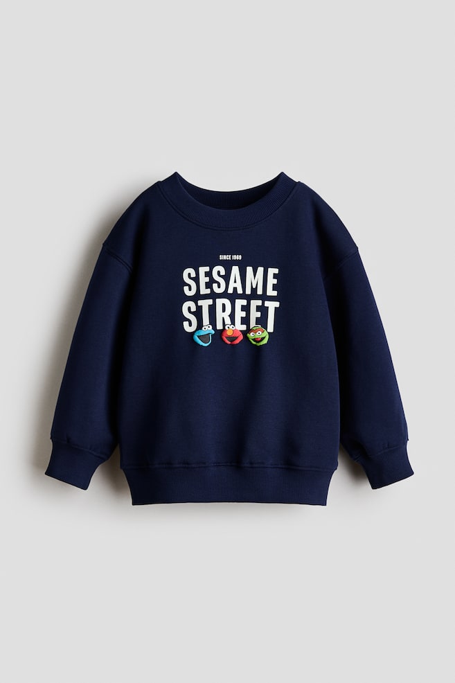 A navy blue sweatshirt is showing its front, displaying a white "SINCE 1969 SESAME STREET" print with three colorful character heads below. It is featuring a round, rib-trimmed neckline, dropped shoulders, long sleeves with ribbed cuffs, and a ribbed hem. - 1