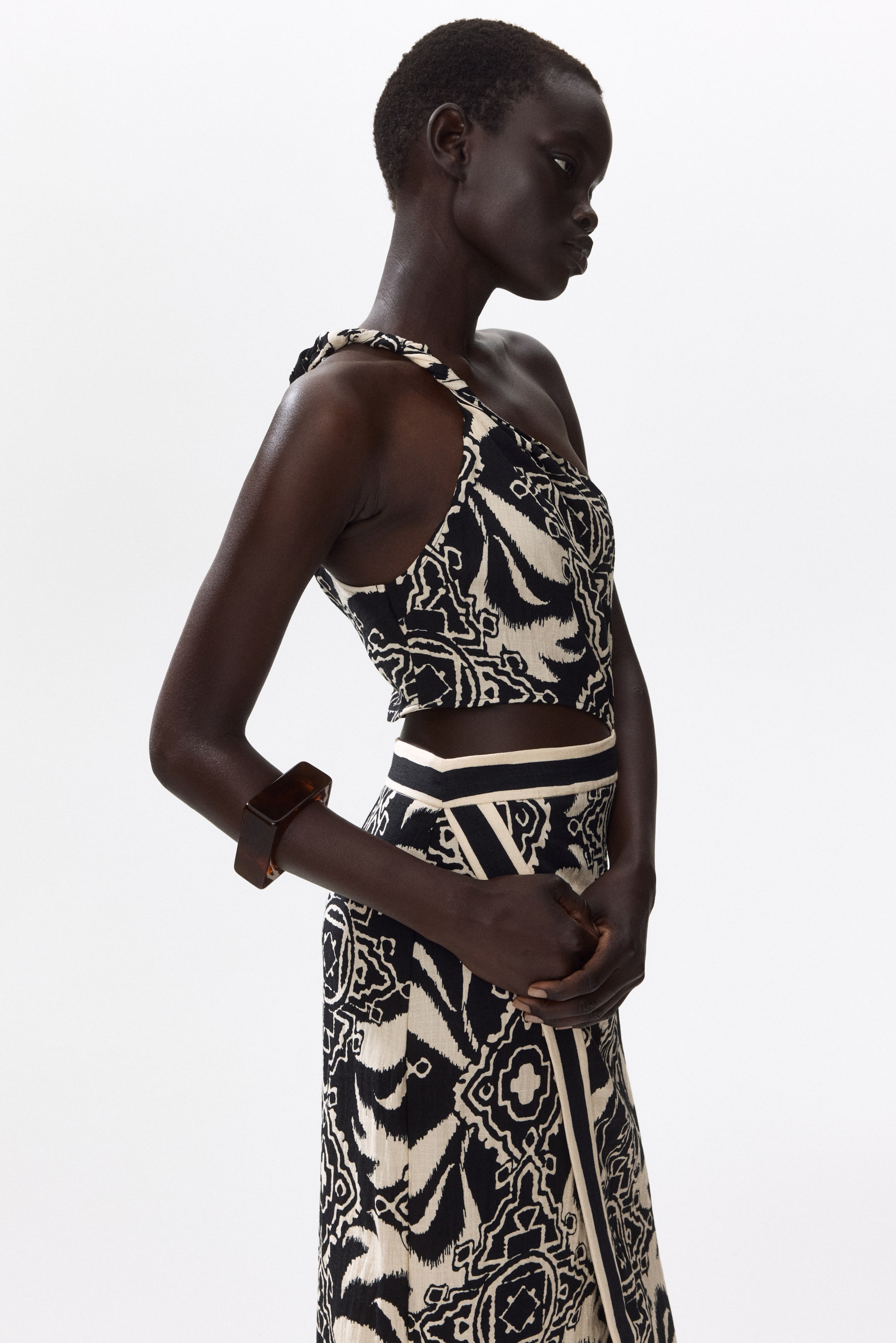 Größeres Bild ansehen: Ein zweiteiliges Set, aus einem leichten Seitenwinkel betrachtet, präsentiert ein asymmetrisches One-Shoulder-Crop-Top mit einem gerafften Träger. Der passende Wickelrock zeigt dasselbe schwarz-cremefarbene abstrakte Muster und kontrastierende, einfarbige Bänder entlang der Taille und der vorderen Öffnung.