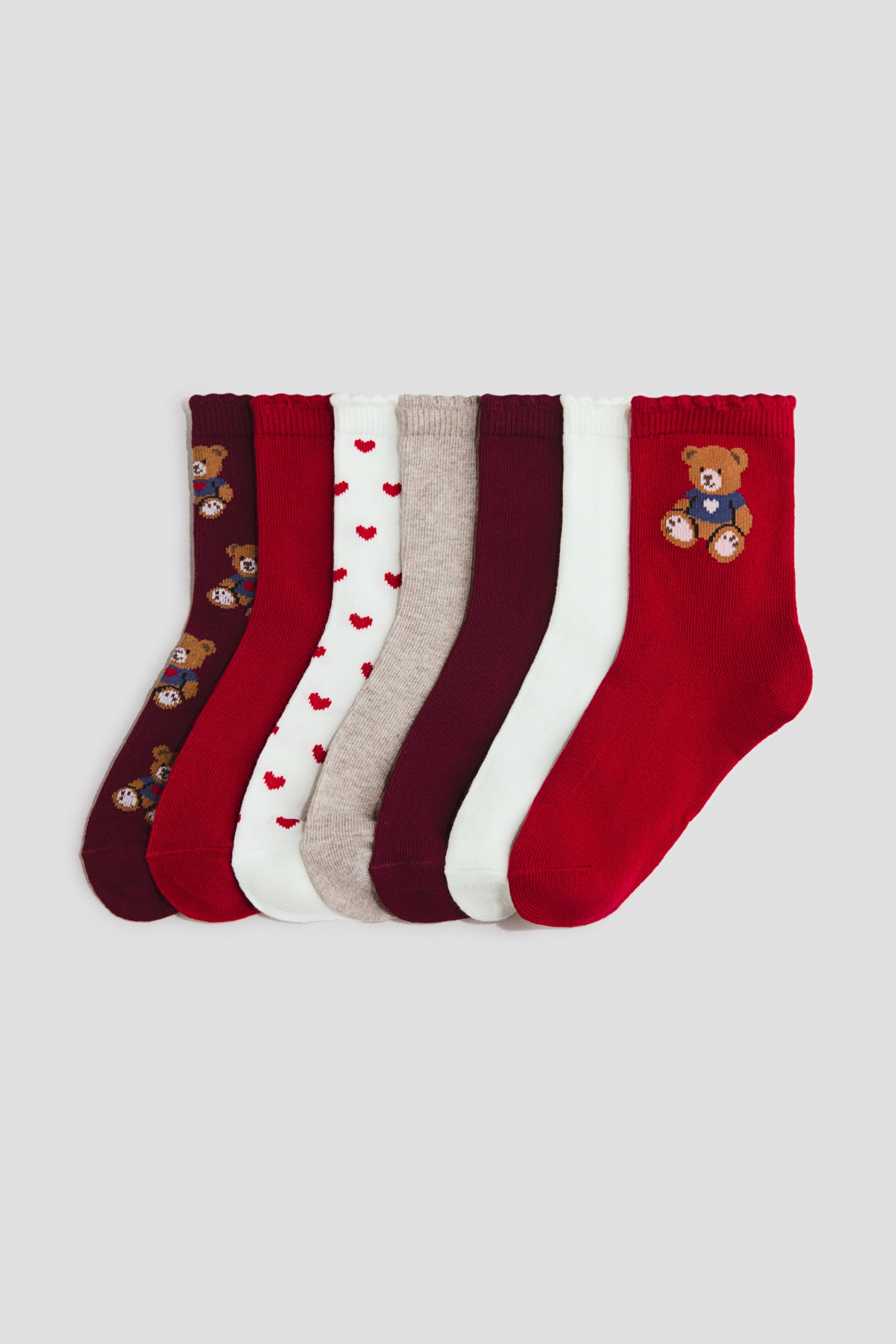 Xem ảnh lớn hơn: Sáu đôi tất dệt kim mịn bằng chất liệu pha cotton mềm mại với cổ tất co giãn đang được trưng bày. Chúng được giới thiệu với nhiều màu sắc và họa tiết khác nhau: một chiếc tất màu đỏ sậm có họa tiết gấu bông lặp lại, một chiếc tất màu đỏ có hình một chú gấu bông lớn gần cổ tất, một chiếc tất màu trắng in họa tiết trái tim đỏ lặp lại, và ba chiếc còn lại có màu trơn đỏ, đỏ sậm và be.