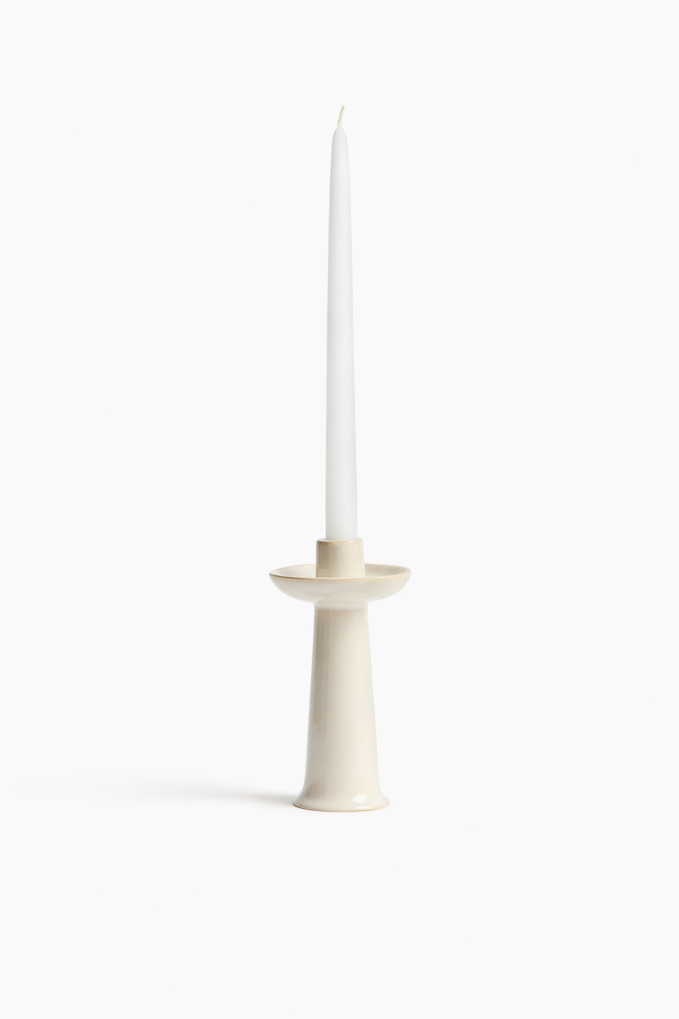 View larger image: This light cream ceramic candlestick showcases a flared base and a tapering body, topped with a dish-shaped surface. It holds a tall, slender white taper candle, extending vertically from its center.