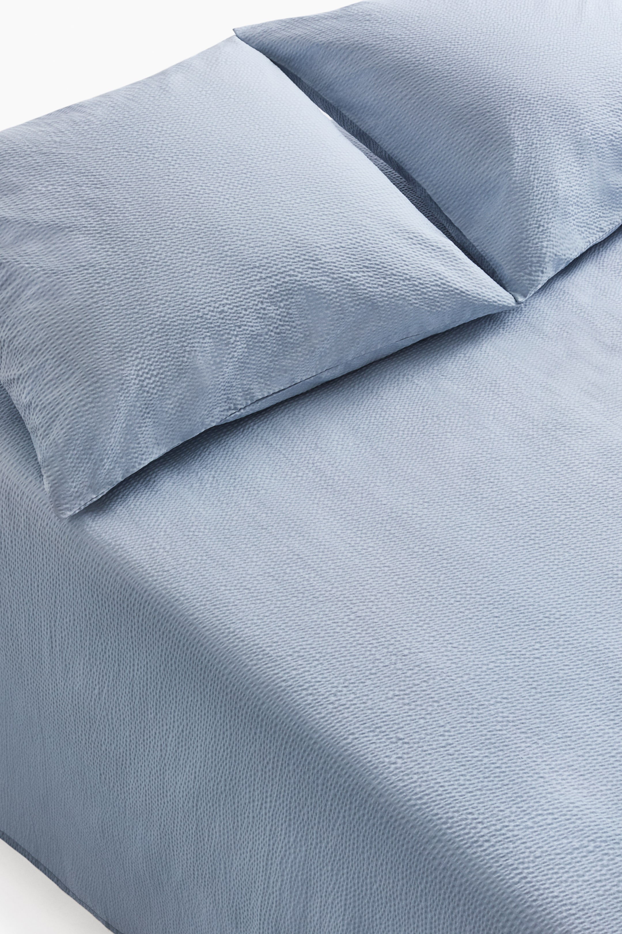 View larger image: A light blue bed linen set is showcased with two pillows neatly arranged at the head of the bed and a matching bedspread elegantly draping over the mattress. The fabric exhibits a subtle, dimpled texture.