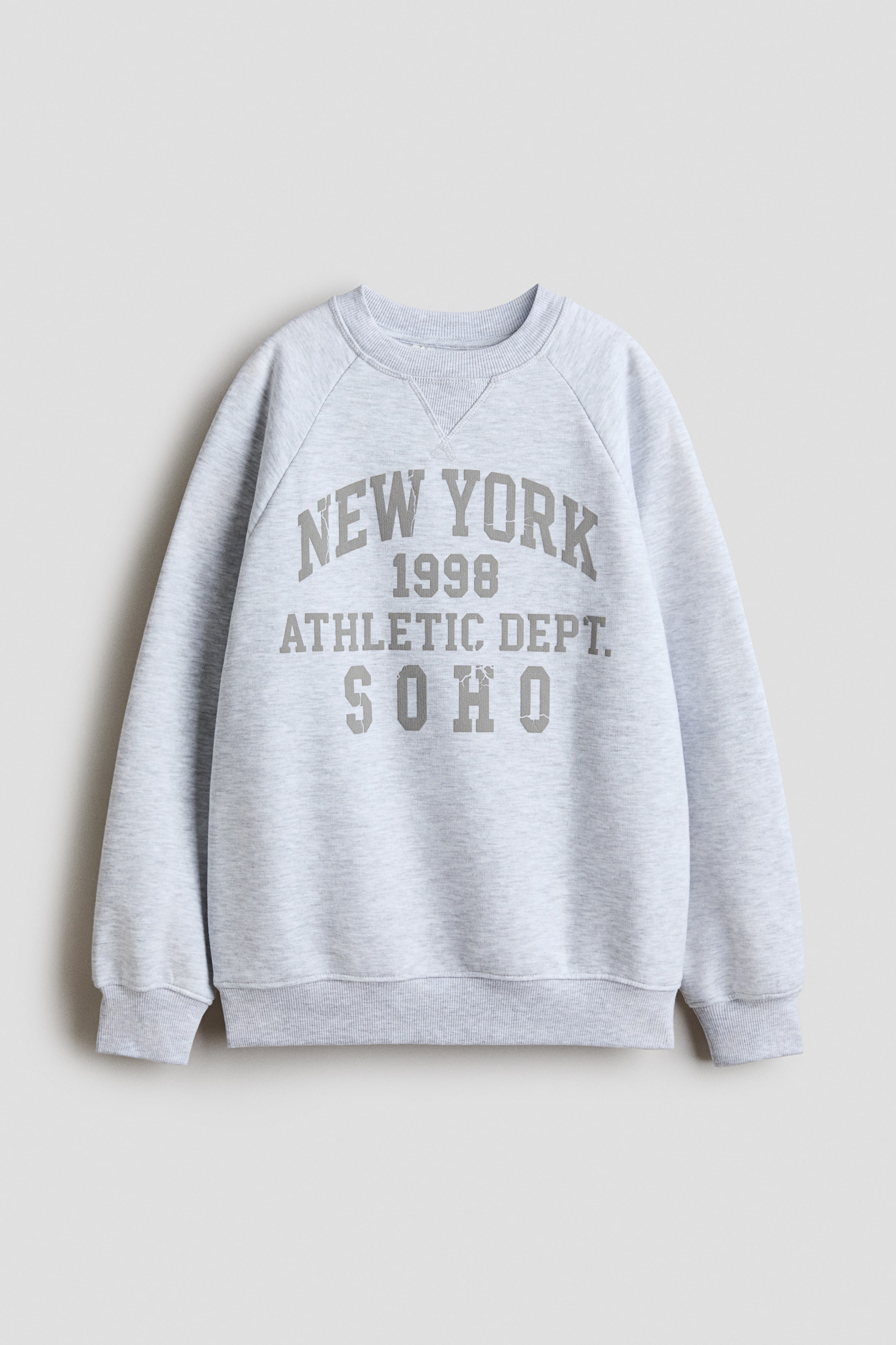 Visa större bild: En sweatshirt med avslappnad passform i ljusgrå melerad är vänd framåt, med rund, ribbad halsringning, långa raglanärmar samt ribbstickade muddar och fåll. Framsidan visar "NEW YORK 1998 ATHLETIC DEPT. SOHO" i grå bokstäver.