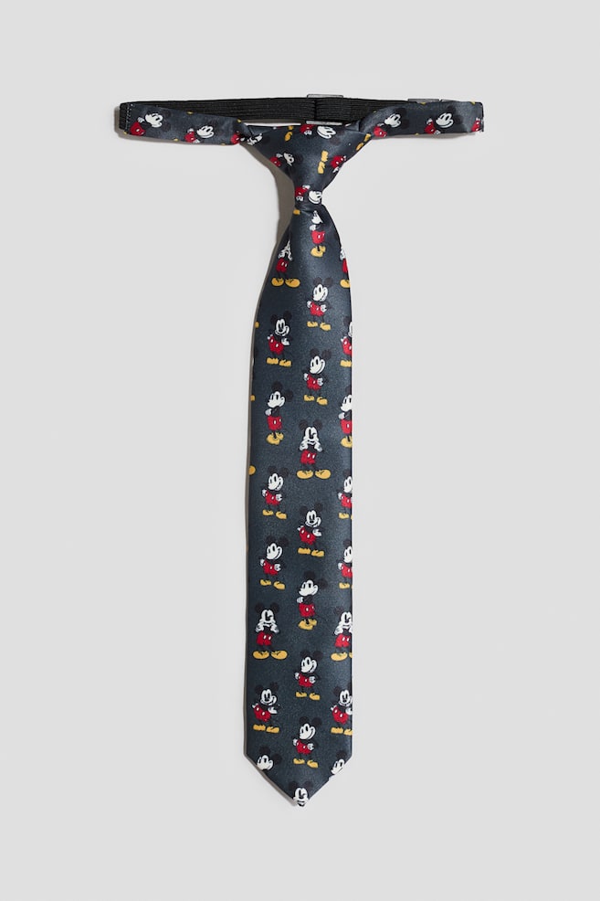 A pre-tied necktie is shown from the front, featuring a repeating Mickey Mouse print on dark grey fabric with a slight sheen. The tie displays a pre-formed knot and a straight blade, with an adjustable black elasticised neck strap extending from the top. - 1