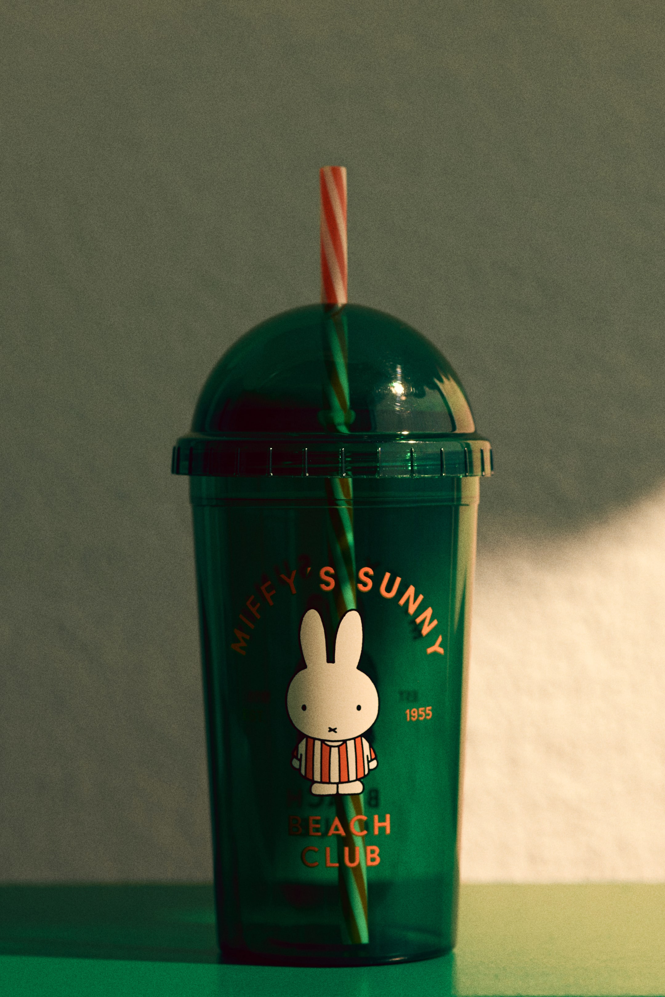 Ver imagem maior: Um copo translúcido verde escuro apresenta uma tampa em forma de cúpula com uma palhinha listrada vermelha e branca a sair dela. Na frente, exibe uma ilustração branca da Miffy a usar uma camisola listrada, com o texto laranja "Miffy's Sunny" acima, "Beach Club" abaixo e "TM 1955" à direita.