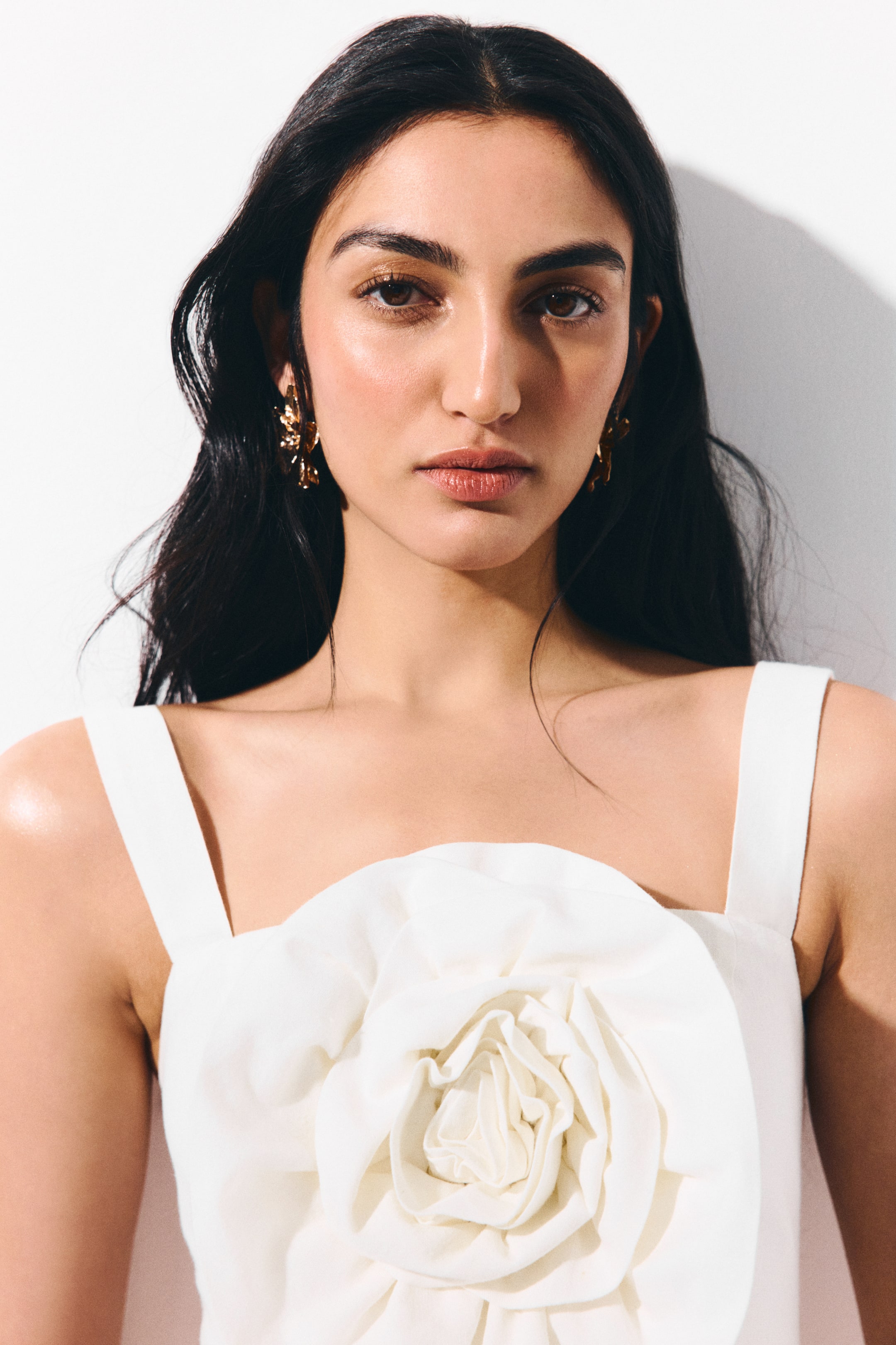 View larger image: A white garment features broad shoulder straps and a striking, three-dimensional fabric floral appliqué centered on the chest. Gold-toned, abstract dangling earrings can be seen on both ears.