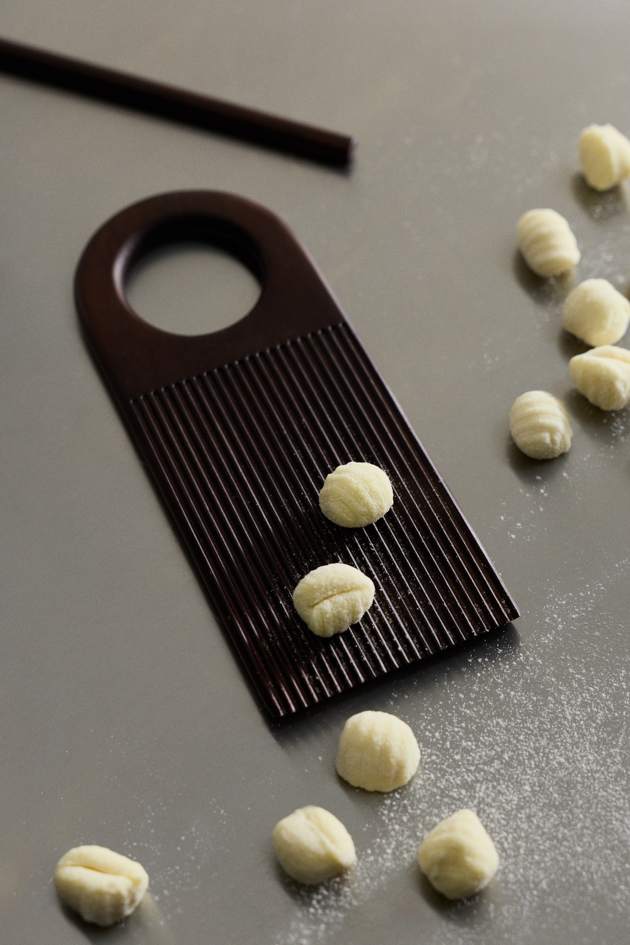 View larger image: A dark brown gnocchi board lies at an angle, showing a rectangular shape with a rounded handle cut-out at one end and a ribbed surface. Several light-yellow gnocchi rest on its ribbed surface, while a matching dark brown rolling rod is also present, used in conjunction with the board.